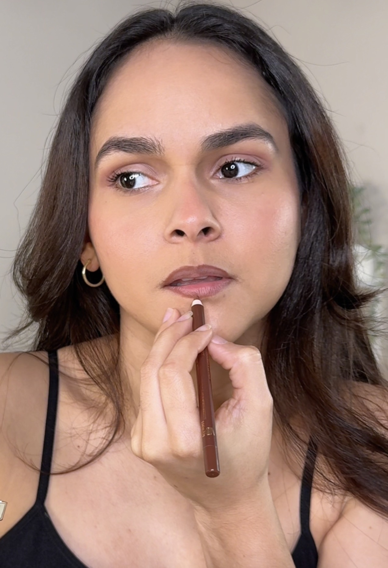 Woman with long dark hair and hoop earrings looks to the side, touching her lips with a makeup brush.