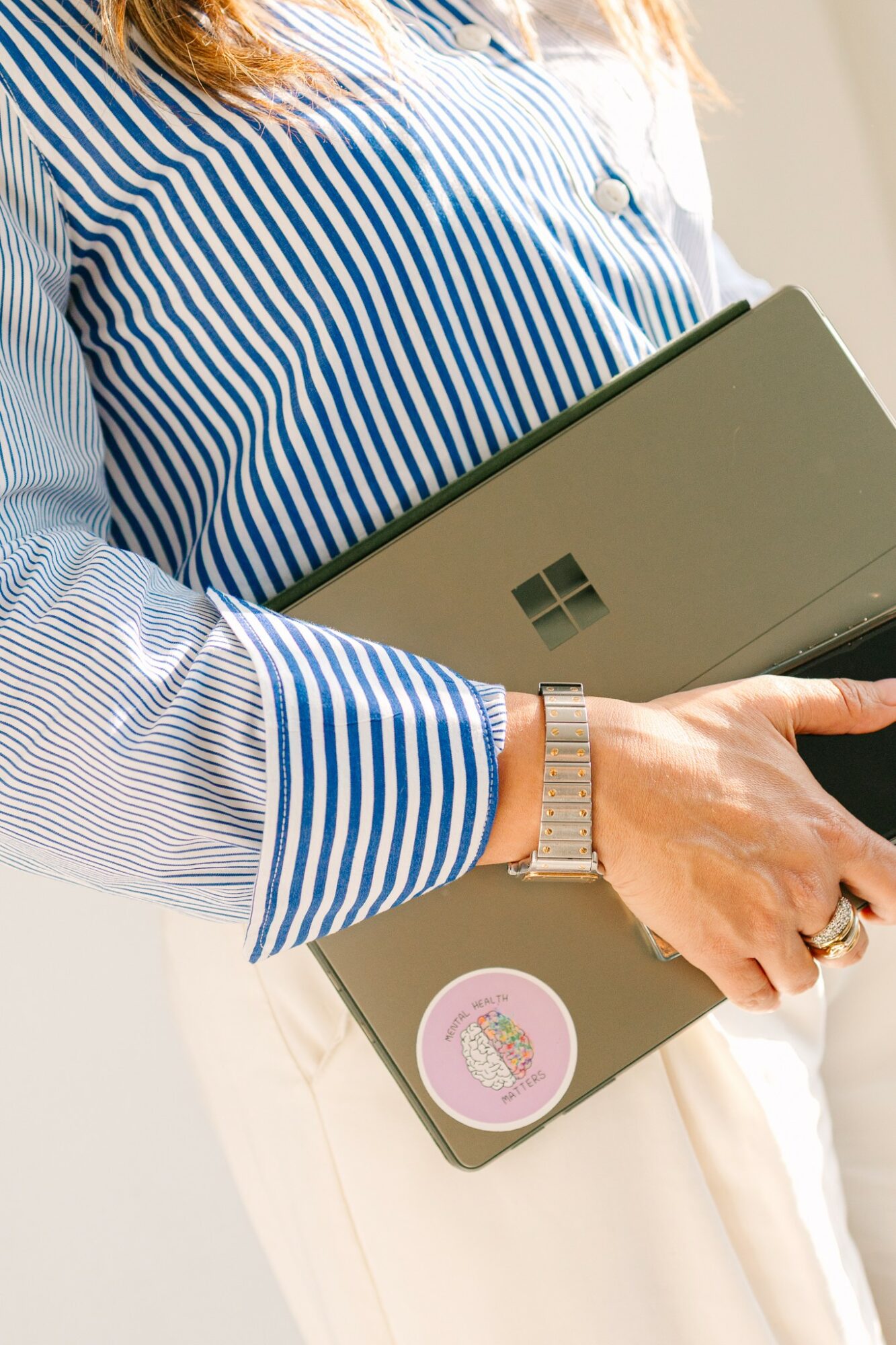 Person holding a laptop with a Windows logo, wearing a striped blue and white shirt and a watch, with a pink sticker on the laptop.