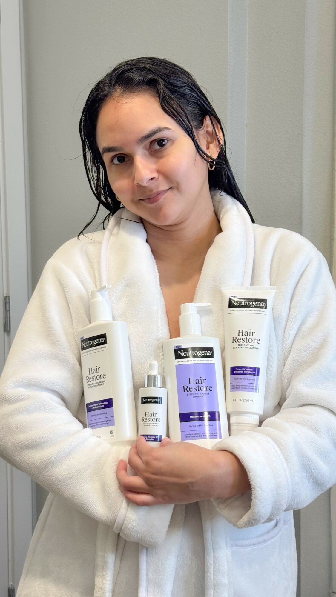 Young woman with dark hair in a white robe holding skincare bottles, standing indoors against a light-colored wall.