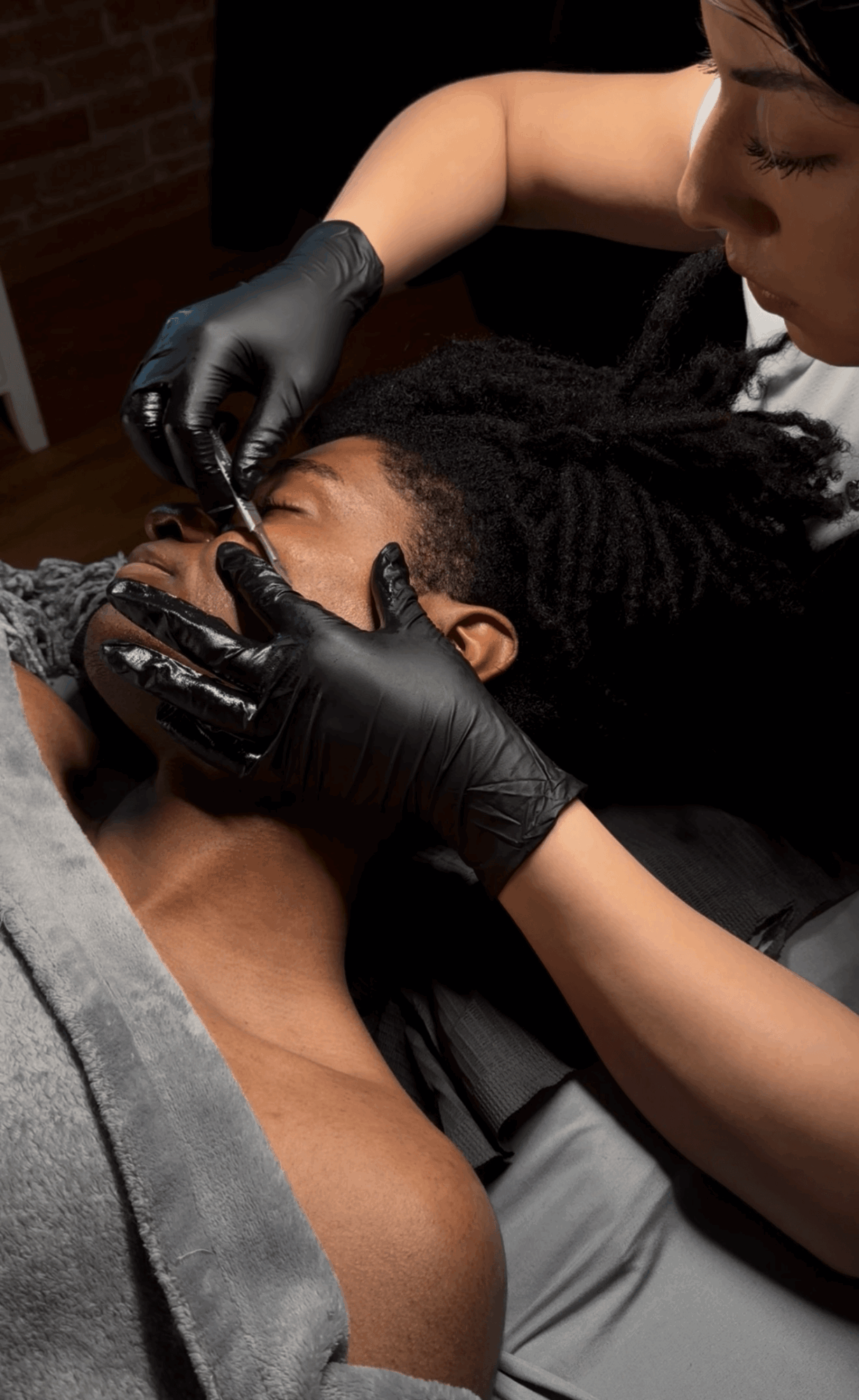 Person receiving a cosmetic procedure on their face, with another person wearing black gloves performing the procedure.