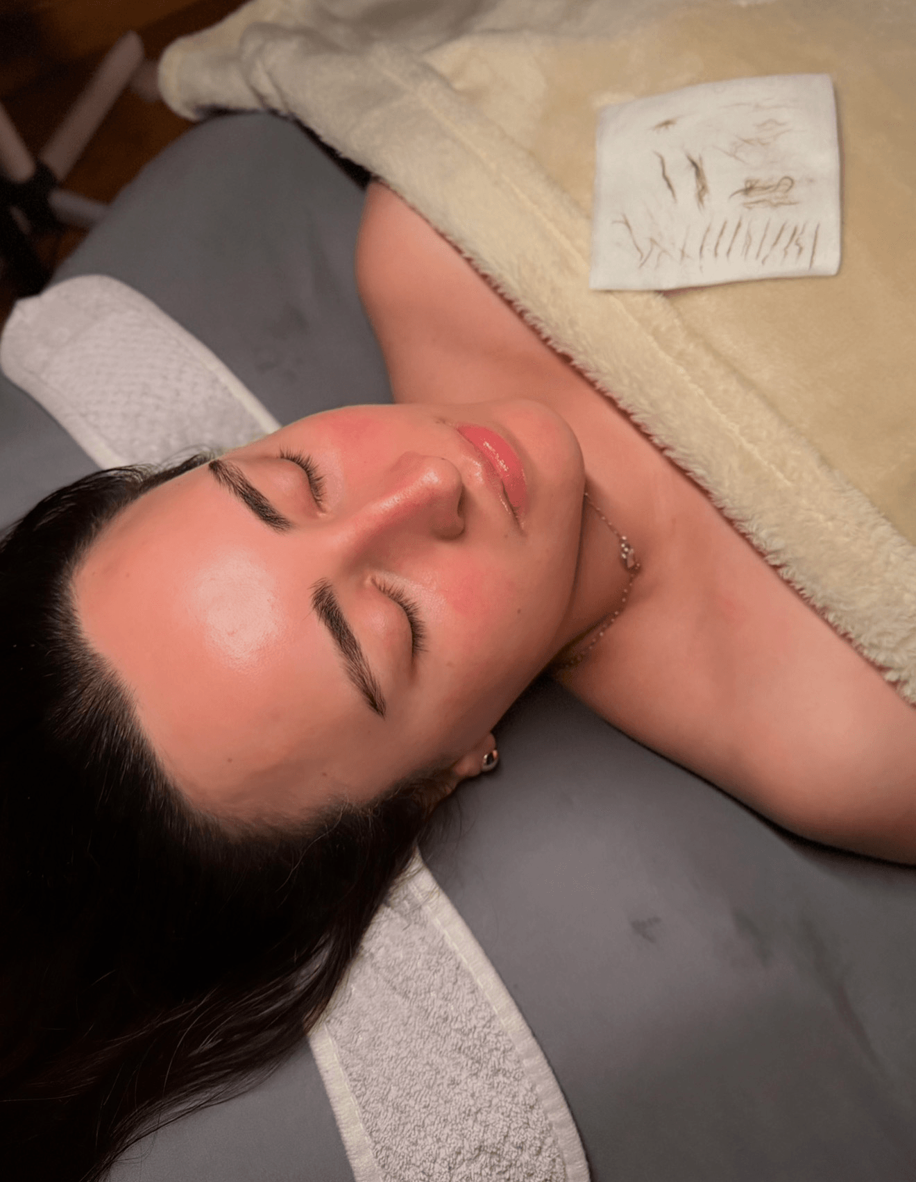 Woman with closed eyes lying on a massage table, covered with a towel, face relaxed, with a paper towel nearby.