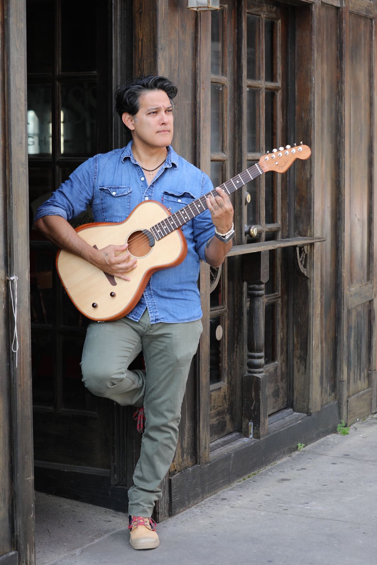 Young man playing guitar outdoors, standing against a wooden fence, wearing a blue shirt and gray pants.
