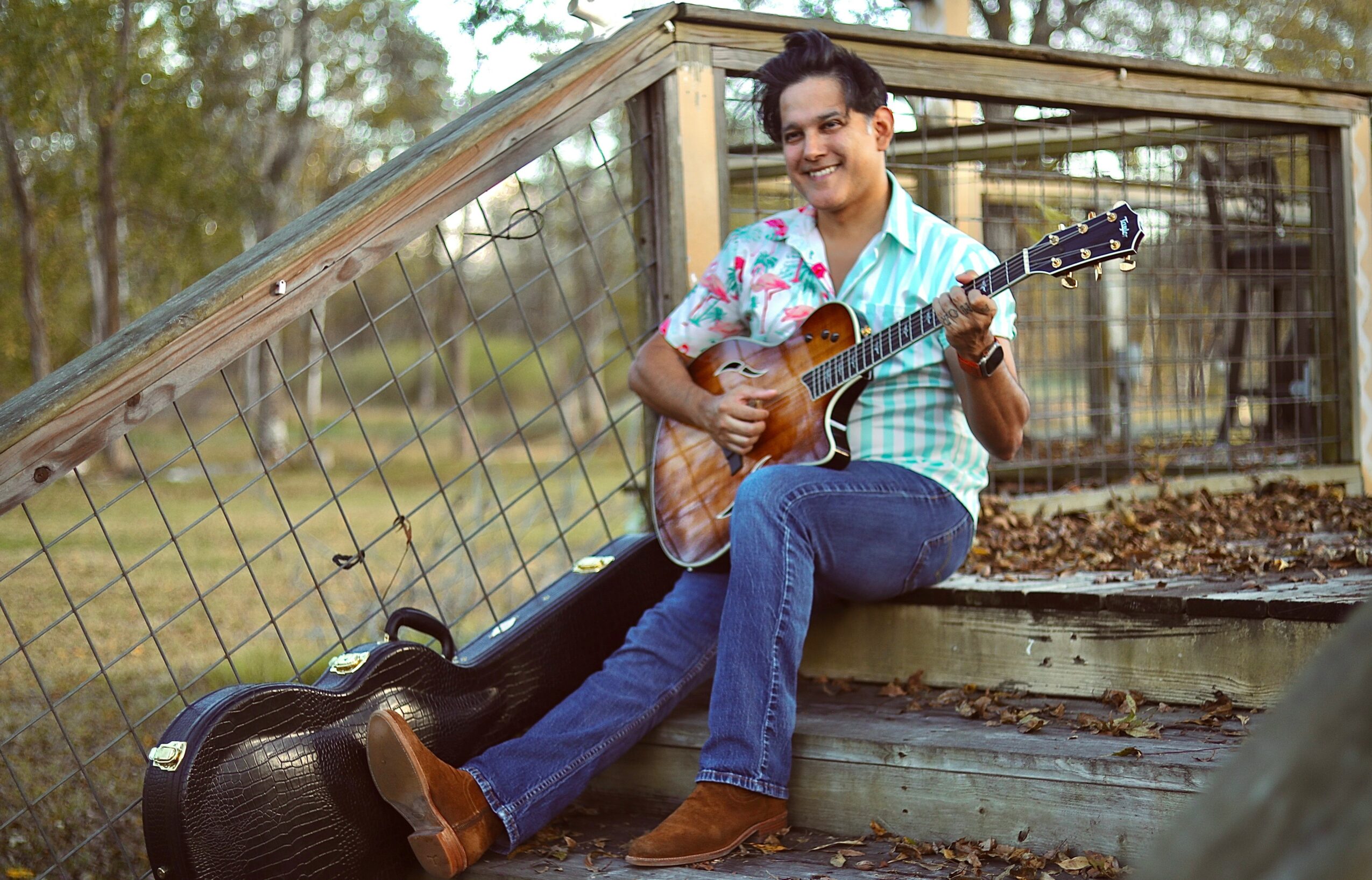 Person sitting on steps playing guitar outdoors, smiling, with a guitar case nearby, in front of a wooden structure.