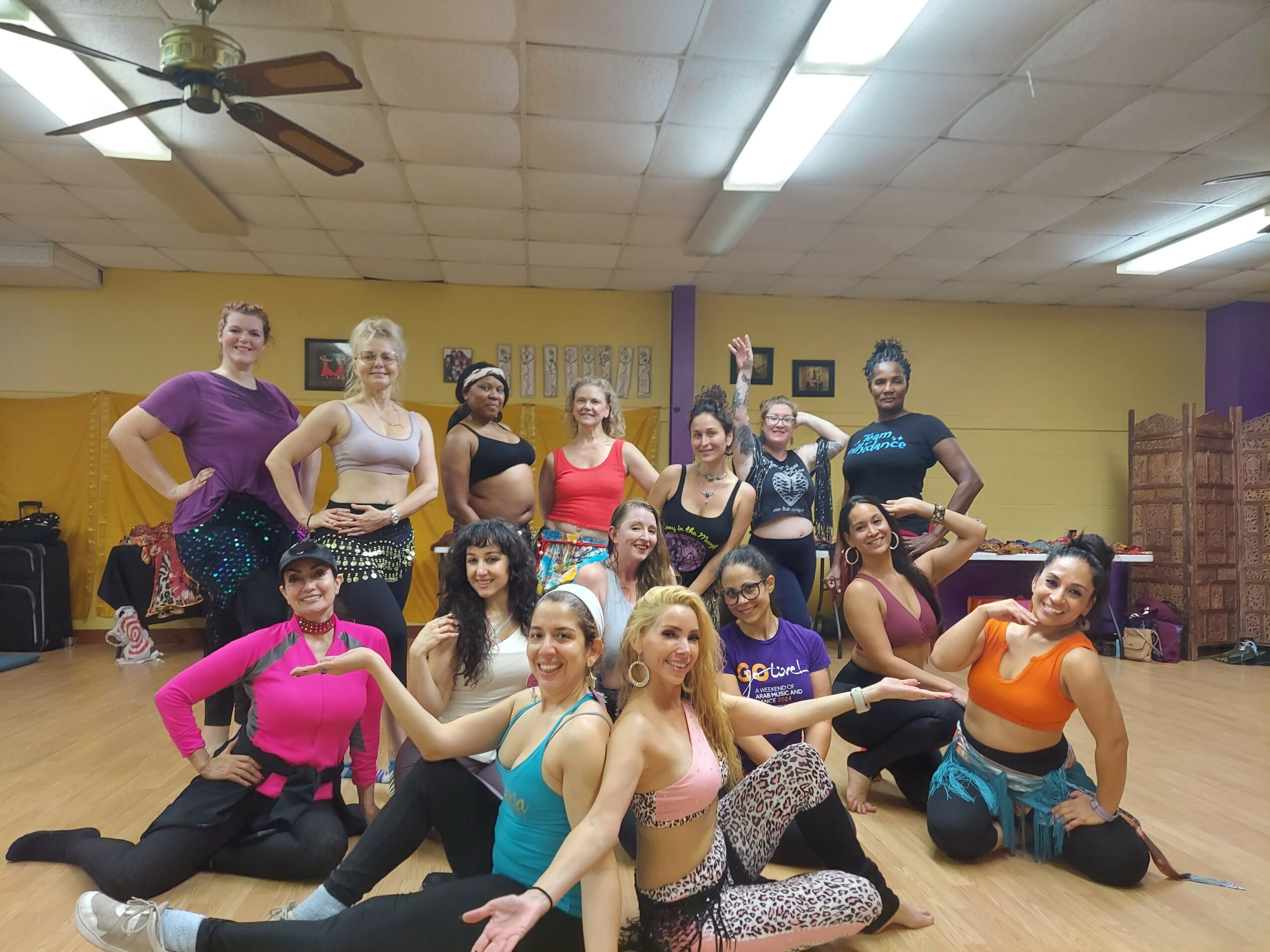 Group of women and girls in dance poses in a studio with yellow walls and wooden floor, some smiling and others striking poses.