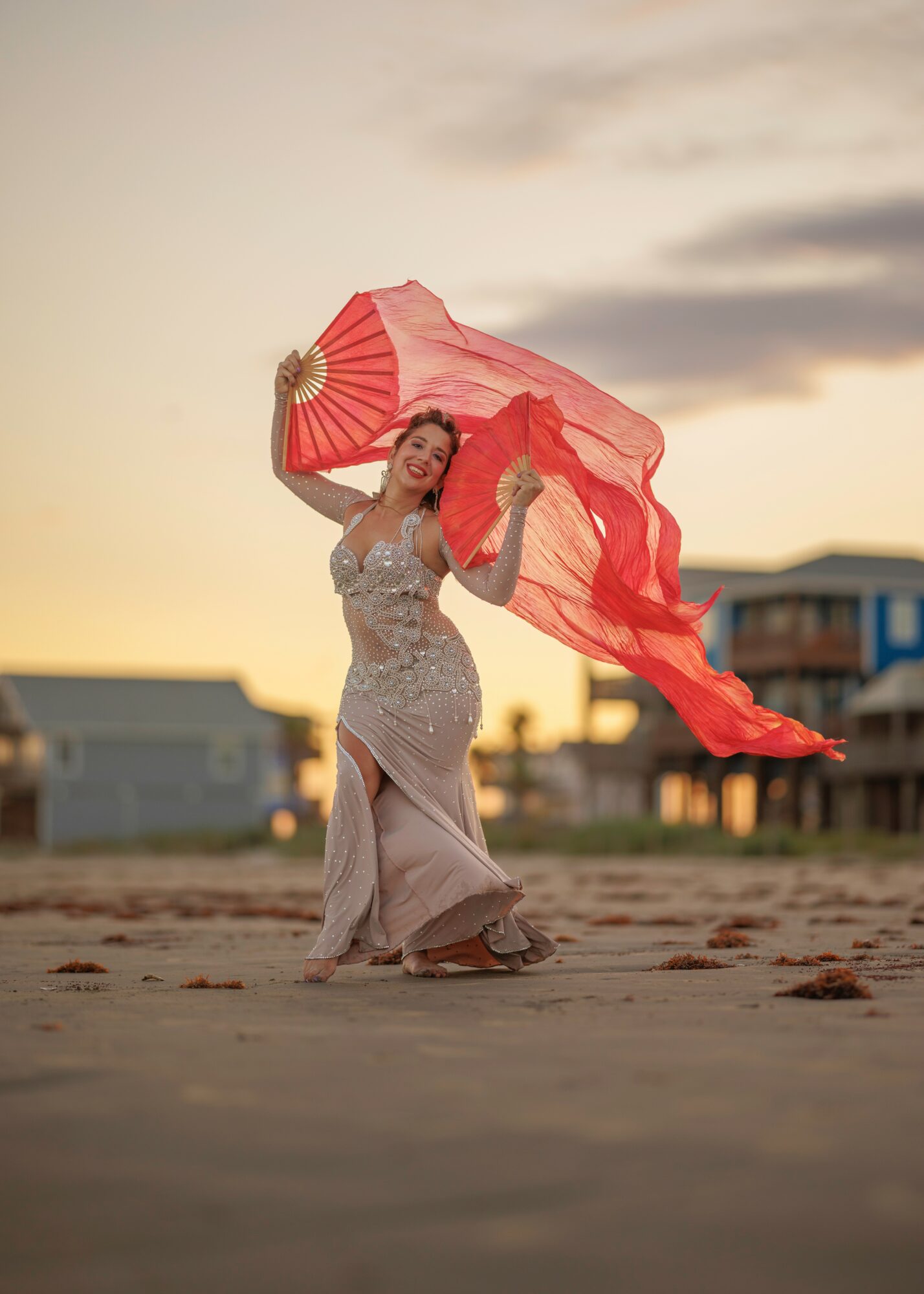 Woman dancing on beach at sunset, holding a flowing pink fabric, wearing a long dress, with houses in background.