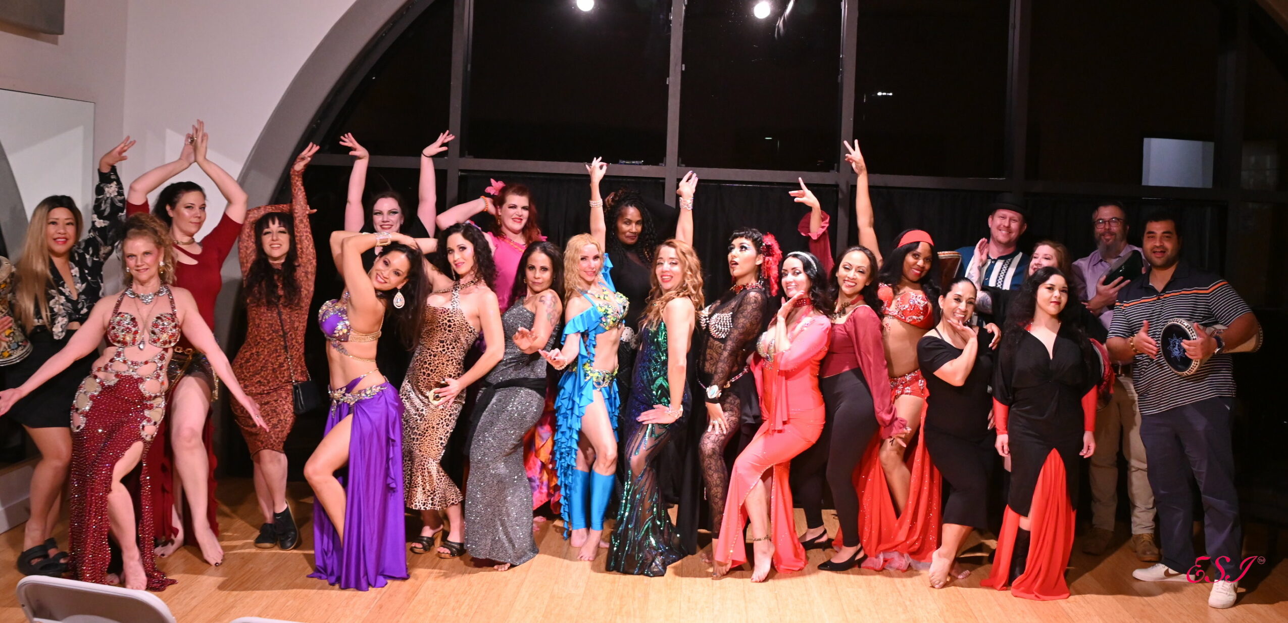 Group of people dancing and posing on stage with a dark background, some holding arms up, wearing colorful costumes.