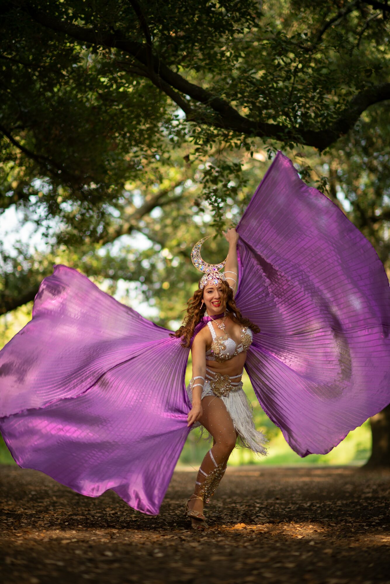 Person in costume with large purple fabric wings outdoors under tree branches, sunlight filtering through leaves.