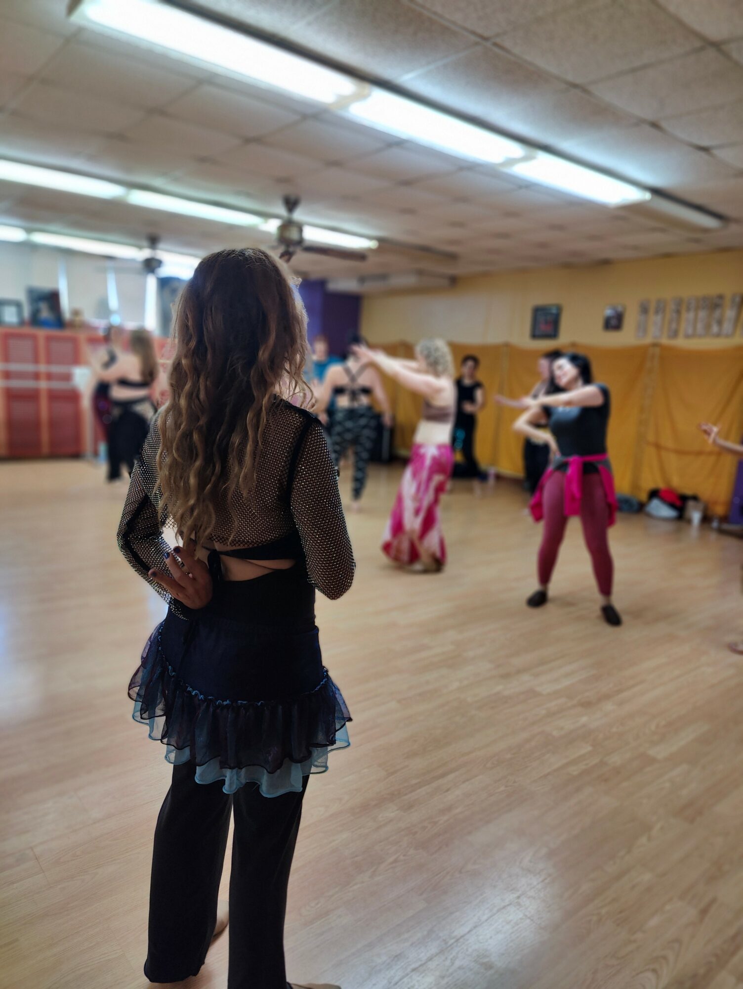 Group of women dancing in a room with wooden floor and yellow walls, some with pink skirts, others with black pants.