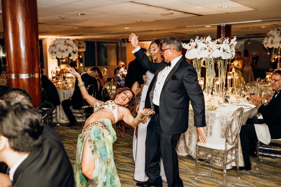 People dancing at a formal event with decorated tables and floral centerpieces, some raising hands and smiling.