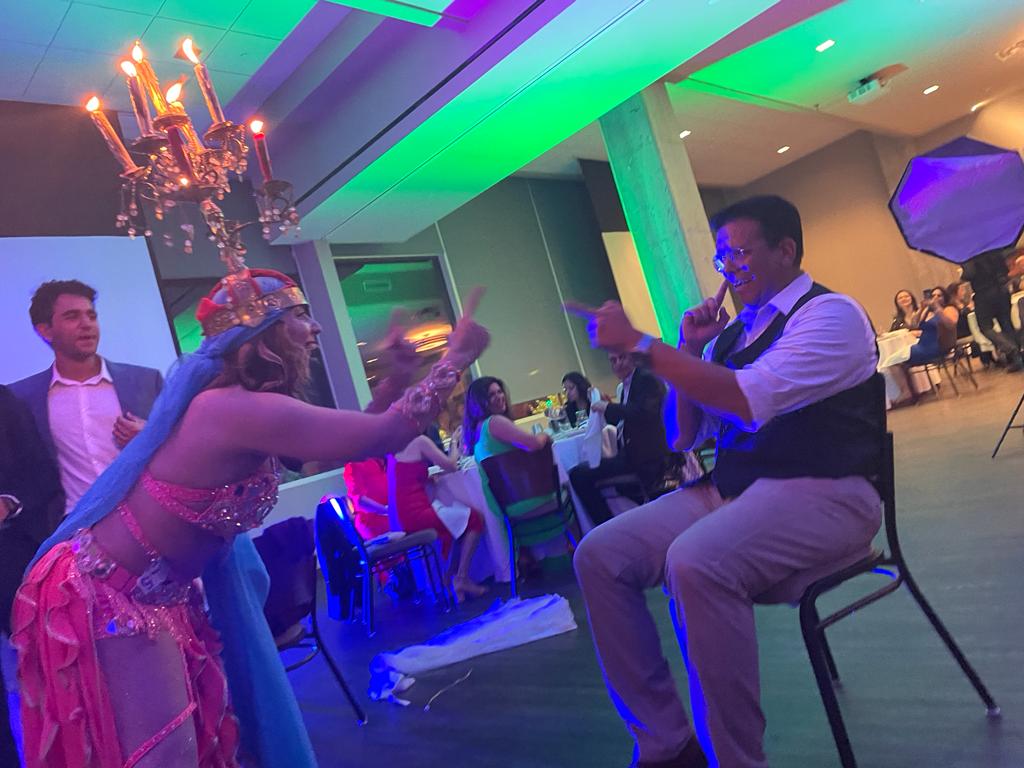 Two people dancing and playing with glow sticks at a colorful indoor event, with a chandelier and seated guests in the background.