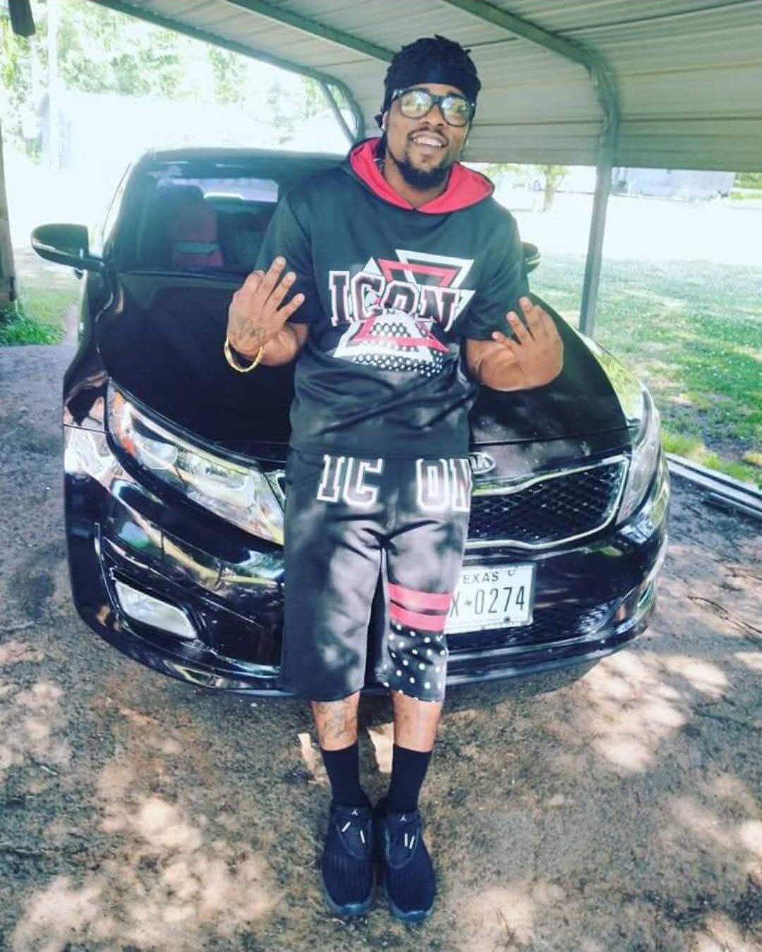 Person standing in front of a black car, smiling, making hand gestures, outdoors with trees and a structure in background.