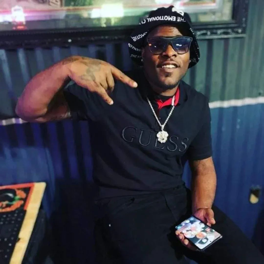 Man wearing sunglasses, a black hat, and a black T-shirt with 'GUESS' logo, pointing to his head, holding a phone, smiling.