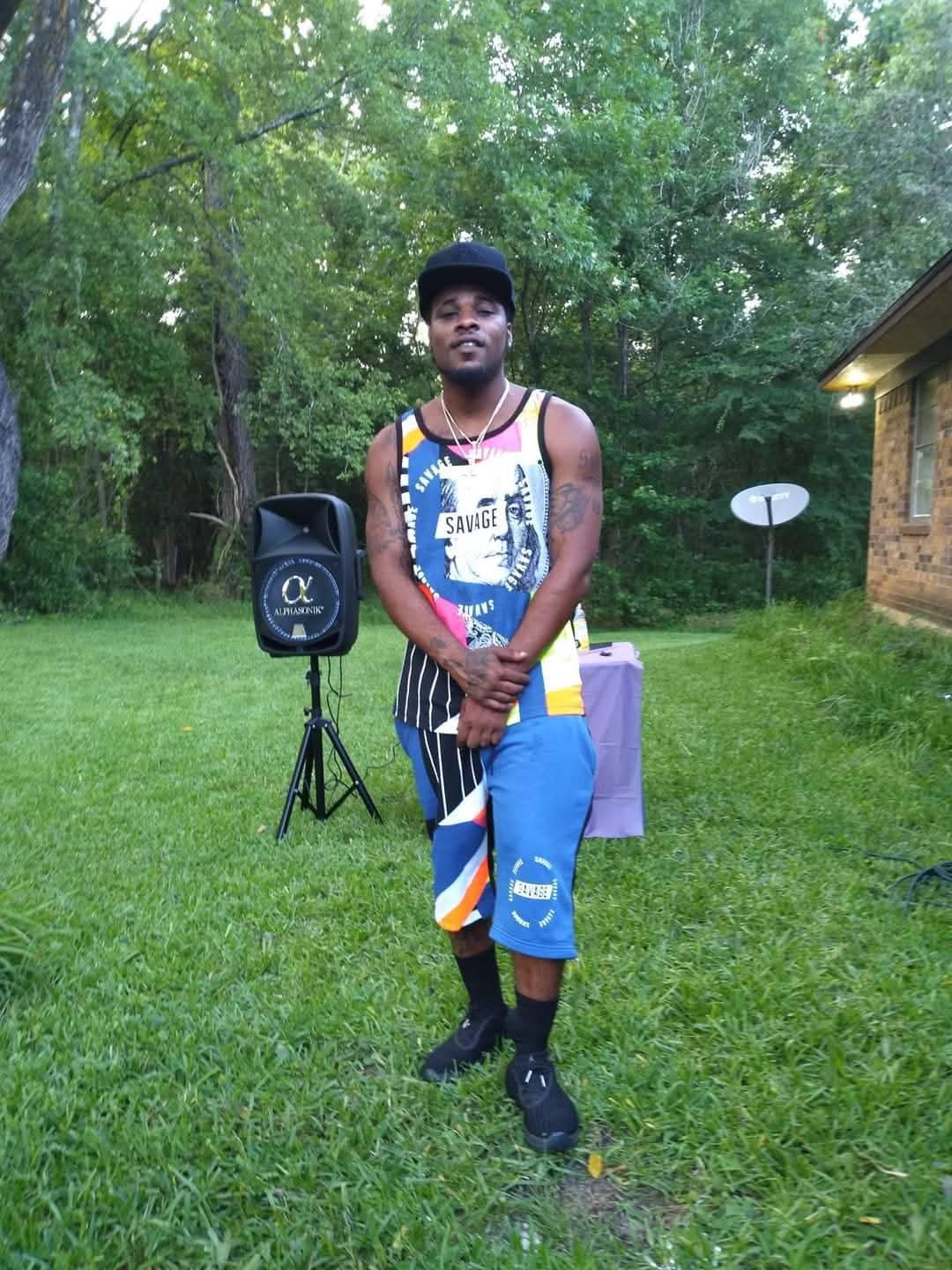Young man standing outdoors on grass, wearing a black hat, colorful sleeveless shirt, striped pants, and sneakers, with trees and a house in the background.