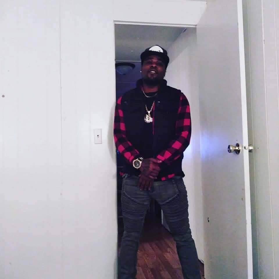 Person standing in doorway wearing a black and red checkered jacket, black shirt, jeans, and accessories, with hands clasped.