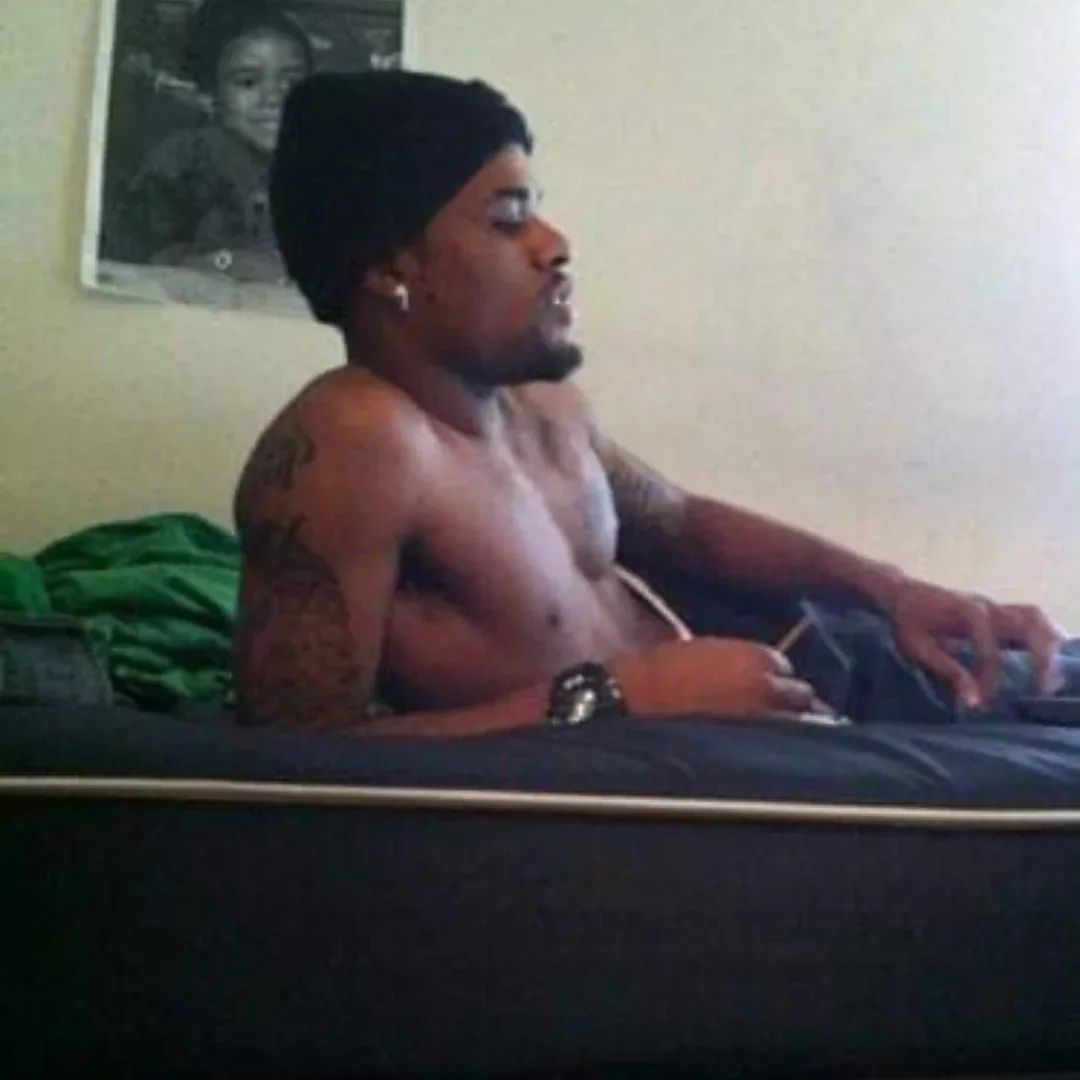 A shirtless man with tattoos and a beanie, sitting on a bed, looking at a device in his hand, with a photo on the wall behind him.