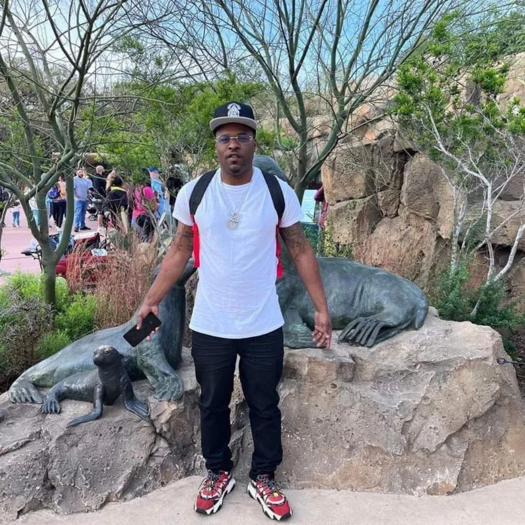 Young man standing outdoors with trees and rocks, holding a phone, surrounded by people, near animal sculptures.