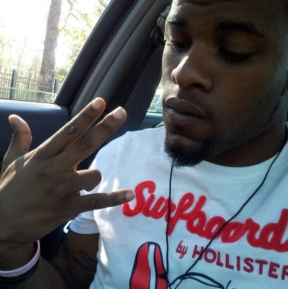 Young man inside a car, holding up three fingers, wearing a white t-shirt with red and blue text, and earphones.