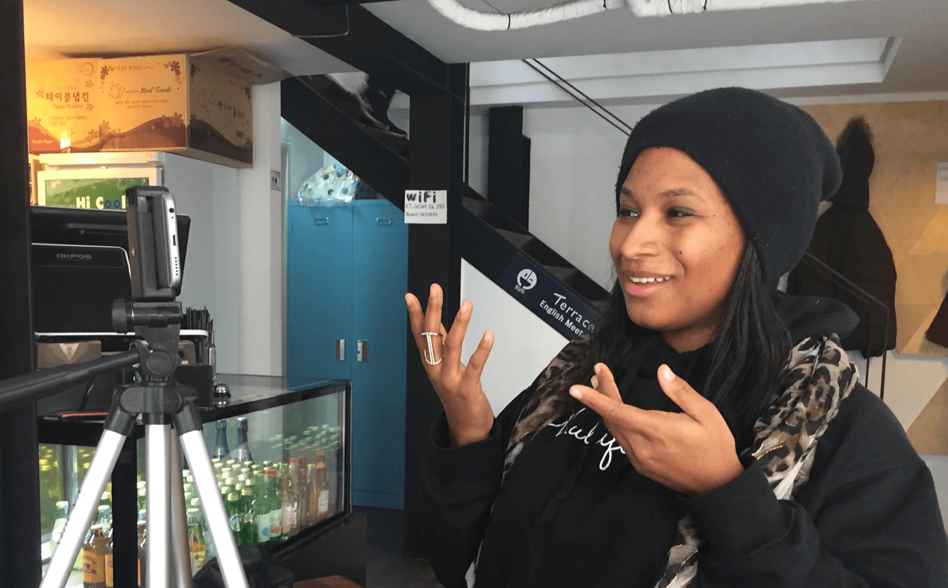 Woman wearing a black beanie and jacket, smiling and gesturing with hands, in an indoor setting with a camera on a tripod.