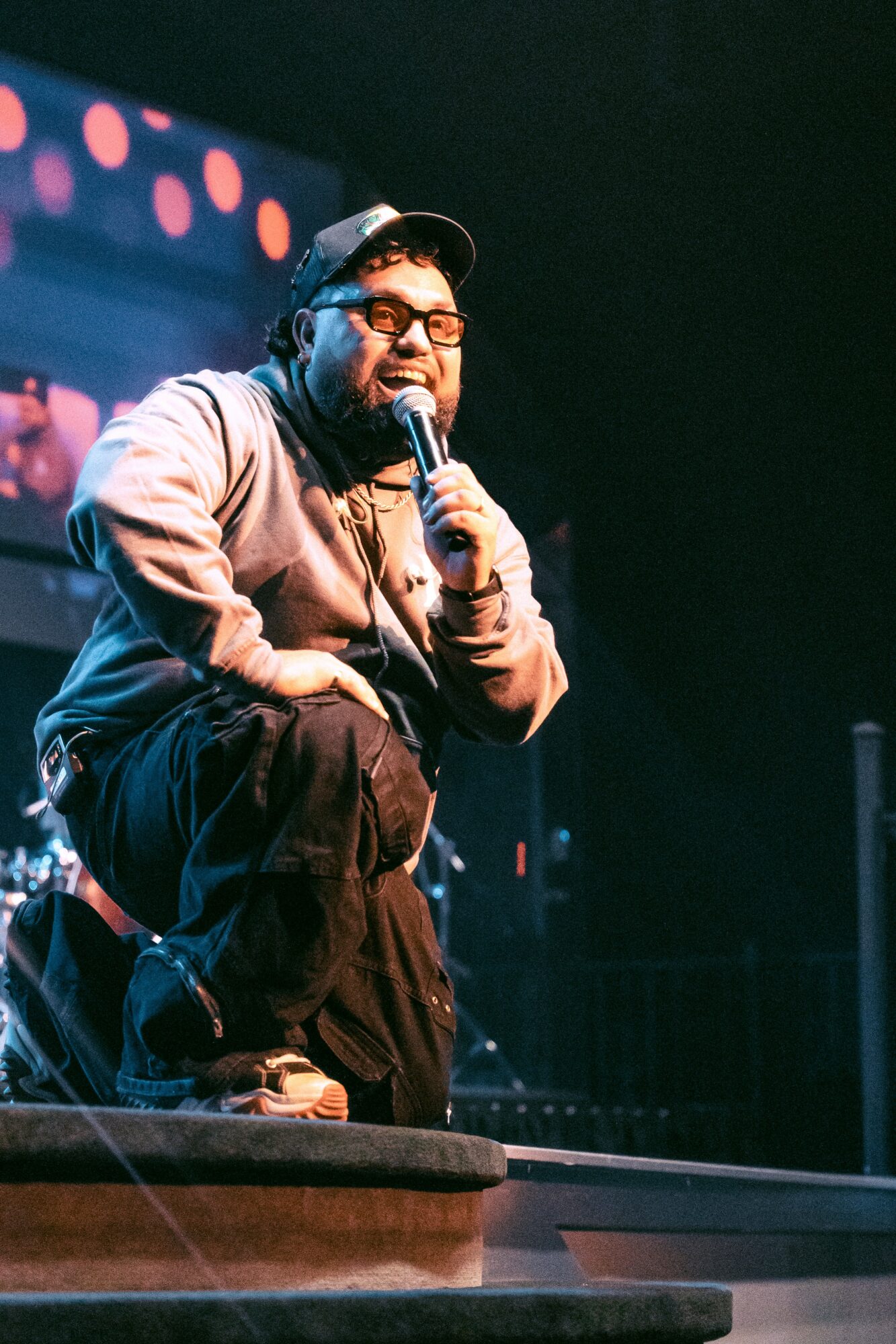 Person crouching on stage, holding microphone, wearing headphones and glasses, with colorful lights in background.