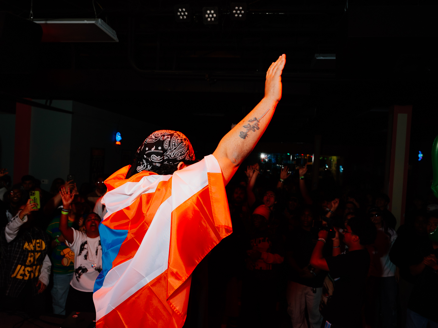 Person with arm raised, wearing a colorful cape, in a dark room with a crowd, facing away from the camera.