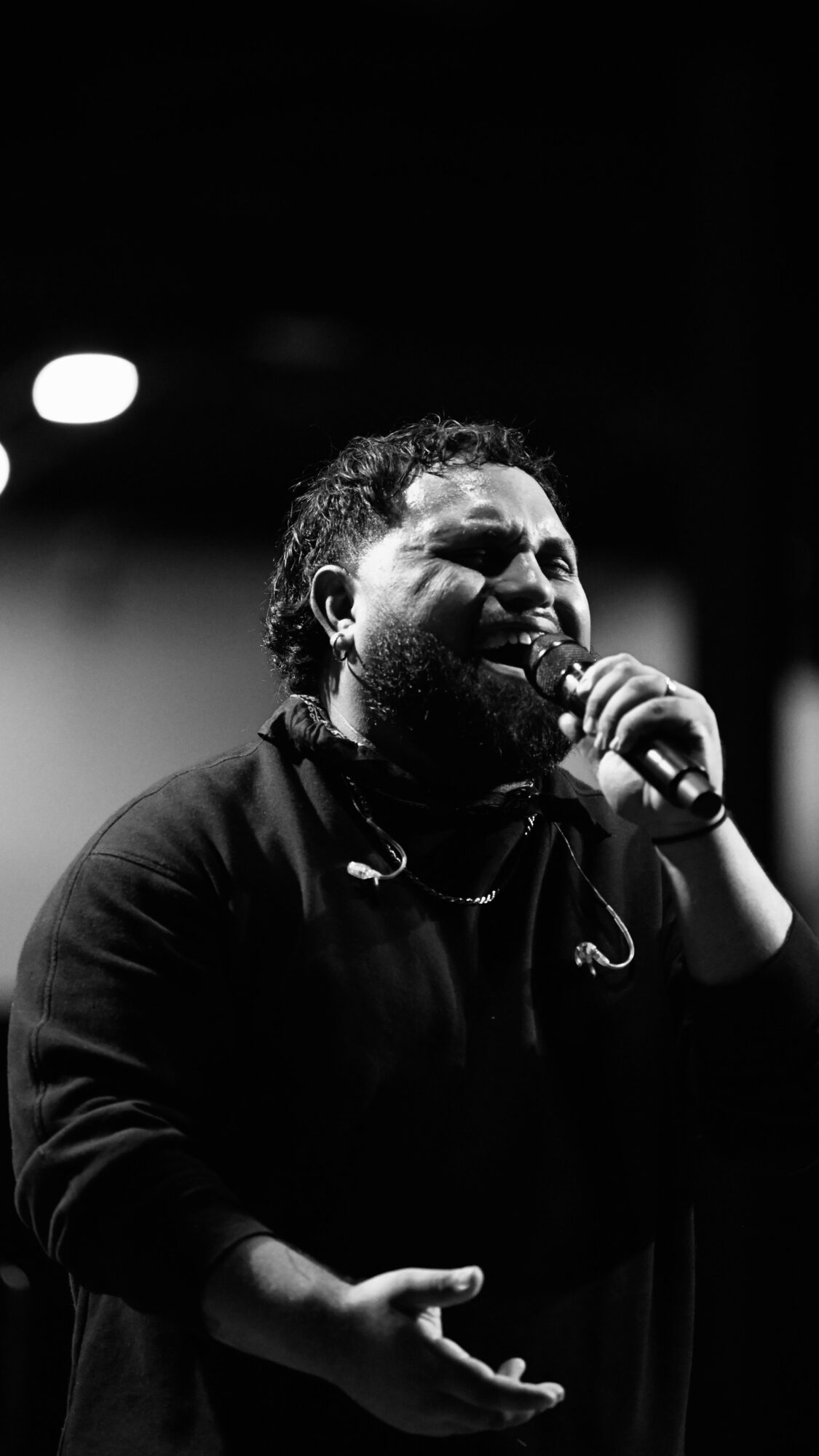 Person singing into a microphone with a dark background, expressive face, and hand gesture, in black and white.