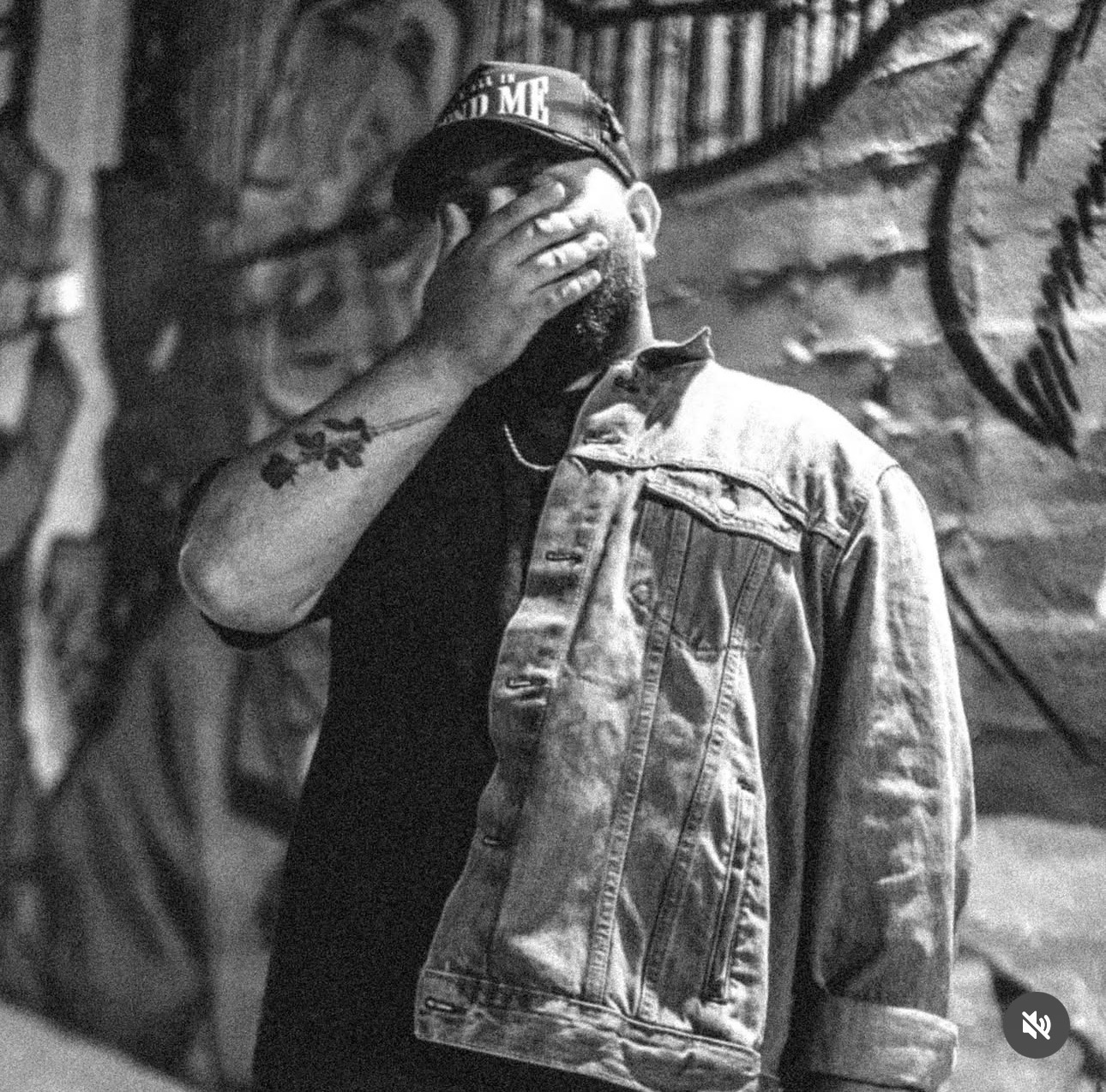 Man with a beard covering his face with his hand, wearing a cap and denim jacket, standing in front of graffiti wall.