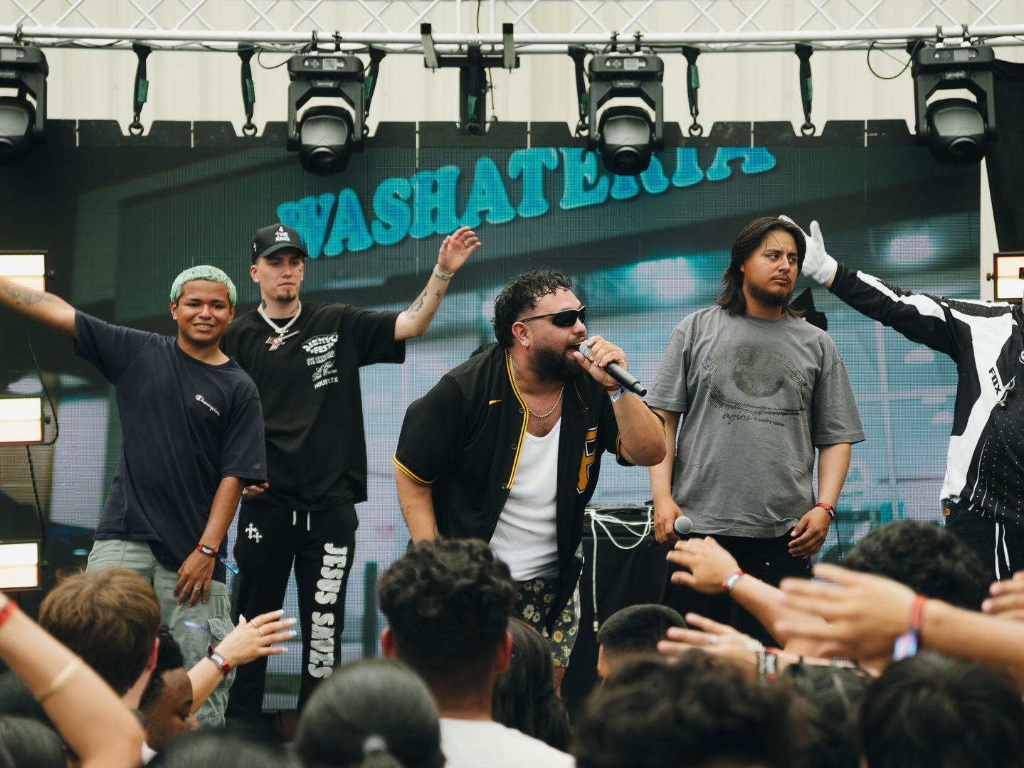 Group of five people performing on stage with a crowd in front, and a sign reading 'YASHATE.COM' in the background.
