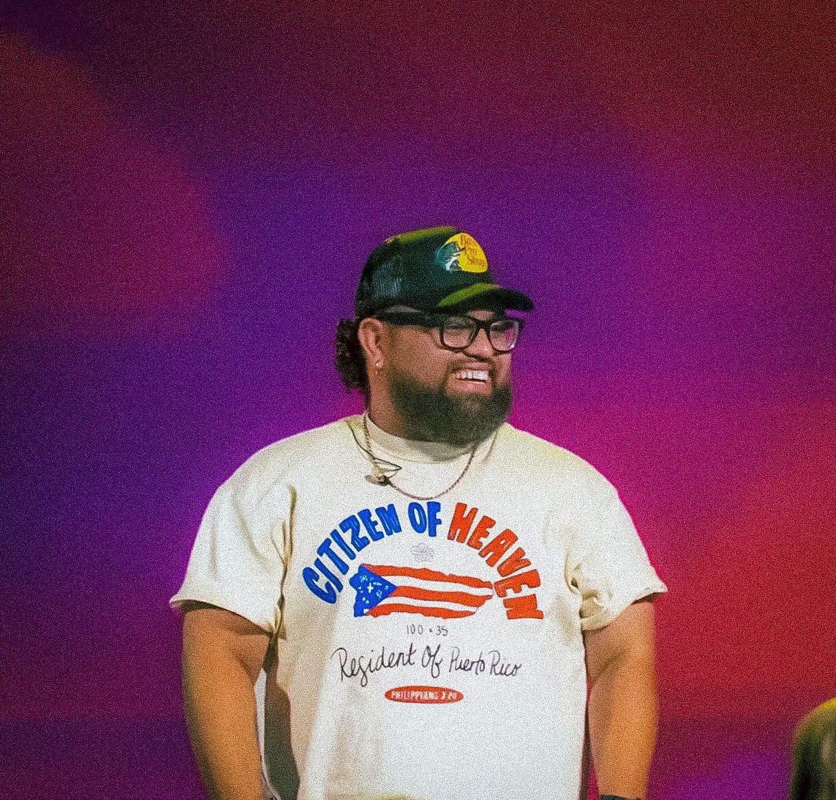 Person with glasses and a beard smiling, wearing a cap and a T-shirt with text, against a purple background.