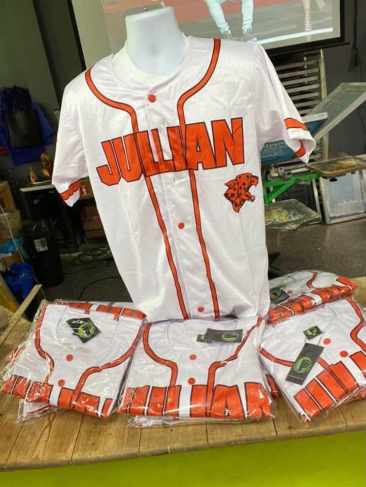White baseball jersey with orange accents, the name 'JULIAN' and a lion logo, displayed on a mannequin, with matching shirts below.