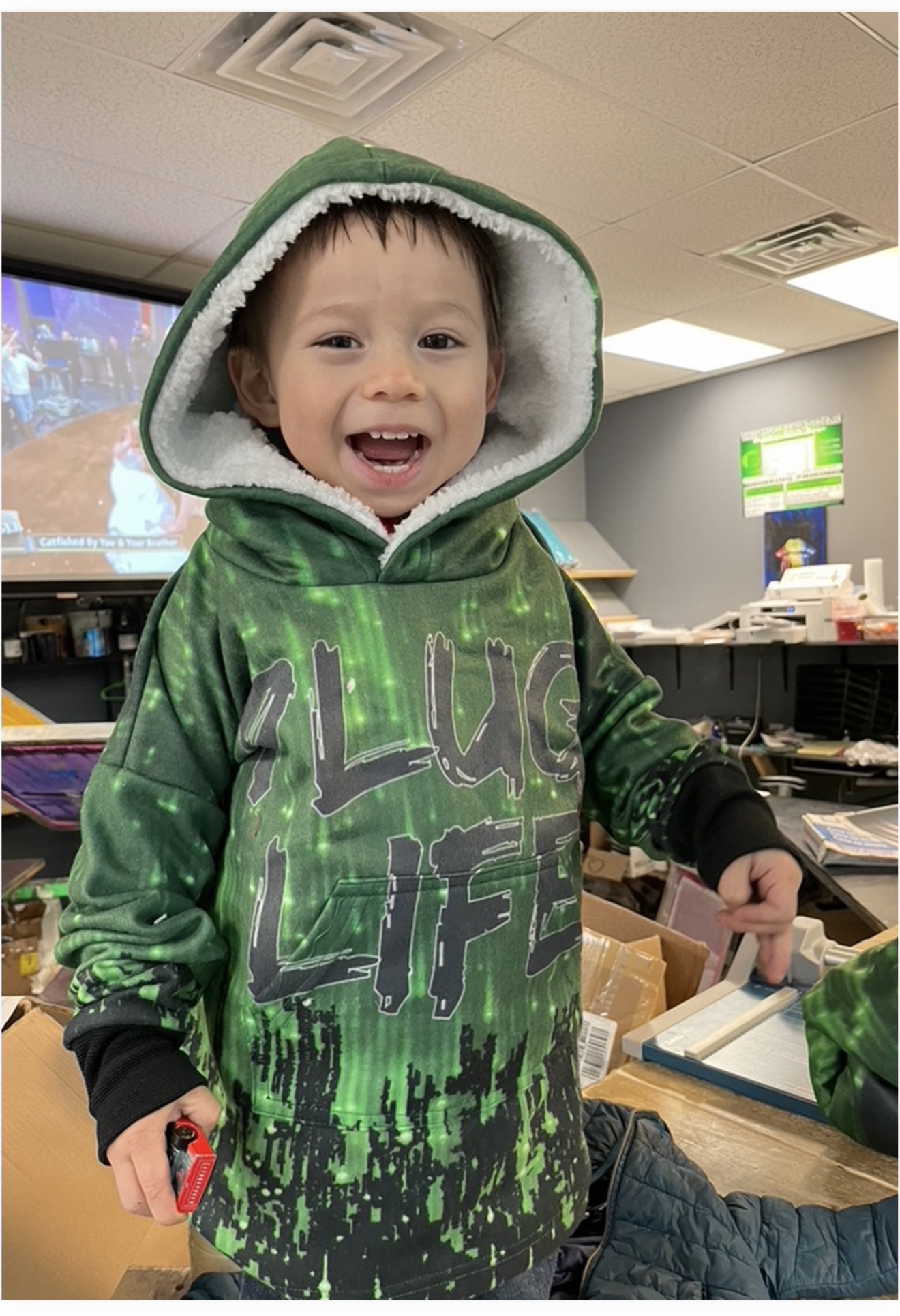 Young boy smiling, wearing a green hoodie with a graphic design, in an indoor setting with desks and a screen.