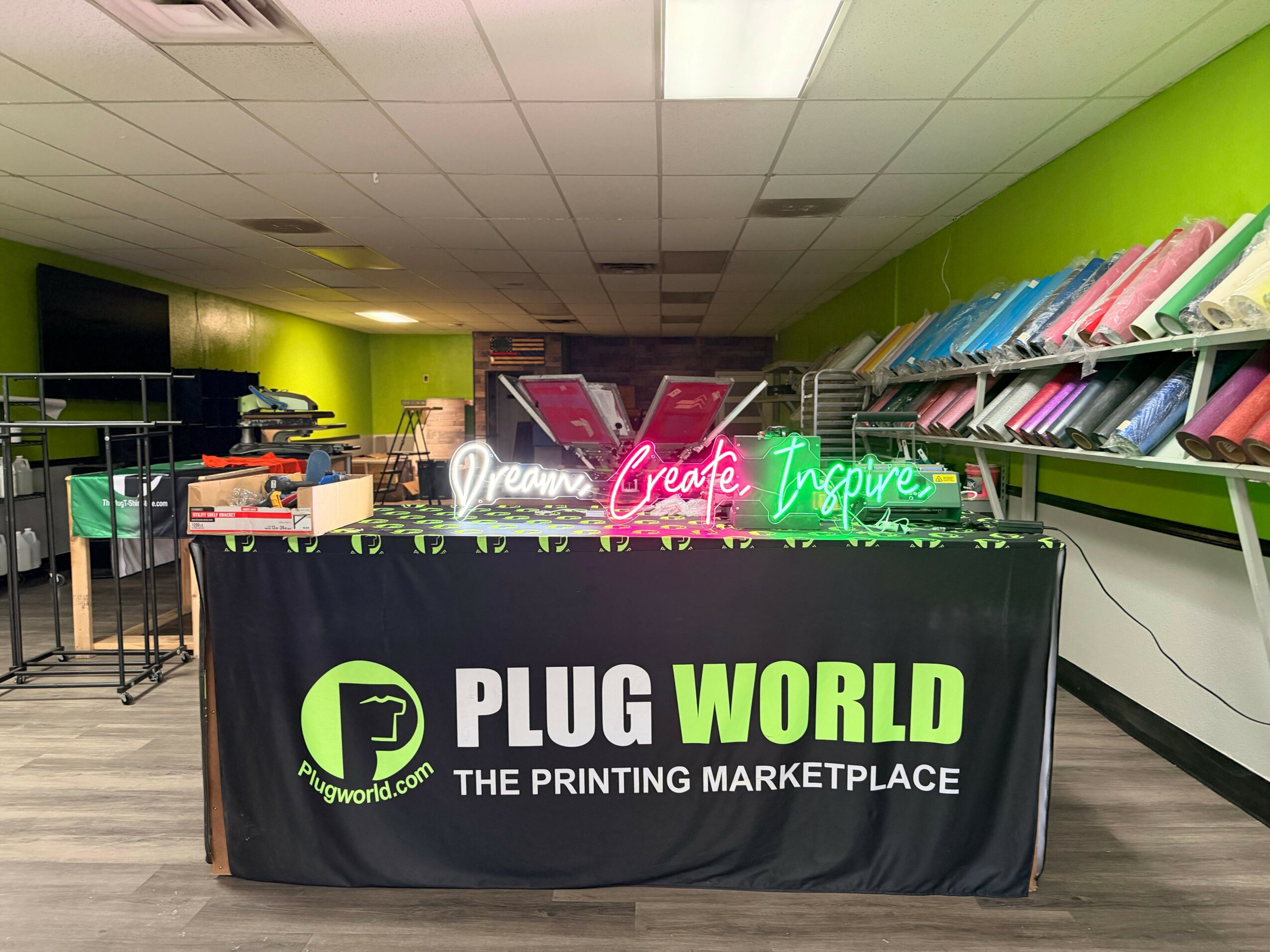 Table with illuminated signs reading 'Dream,' 'Create,' and 'Inspire' in front of a green wall and shelves with colorful paper.