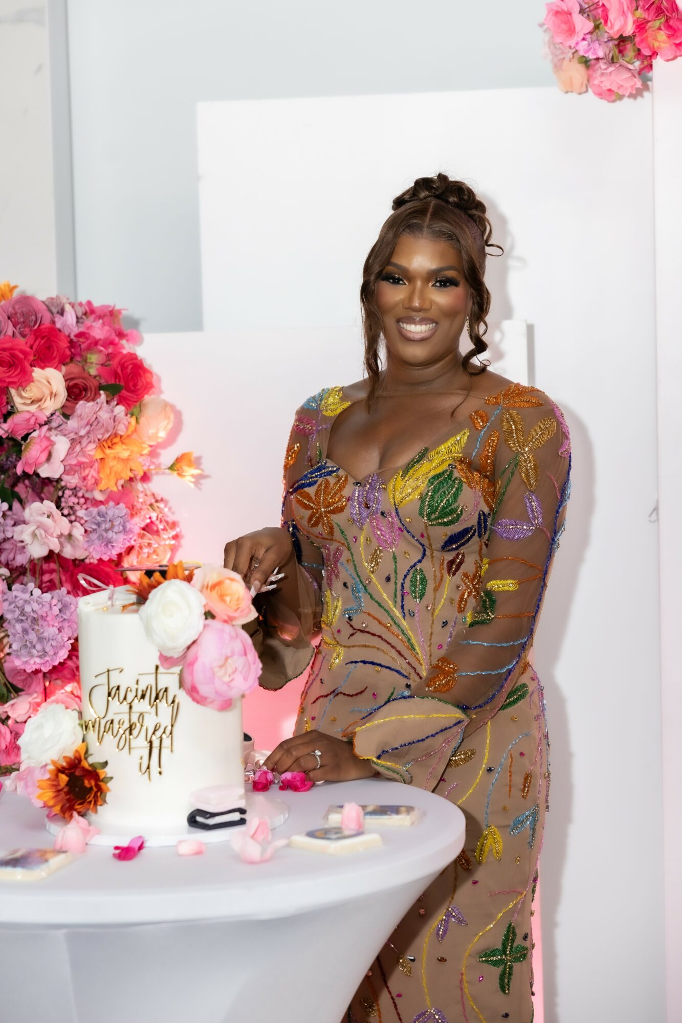 Smiling woman in colorful dress stands near a table with pink flowers and a gift bag, indoors with white background.
