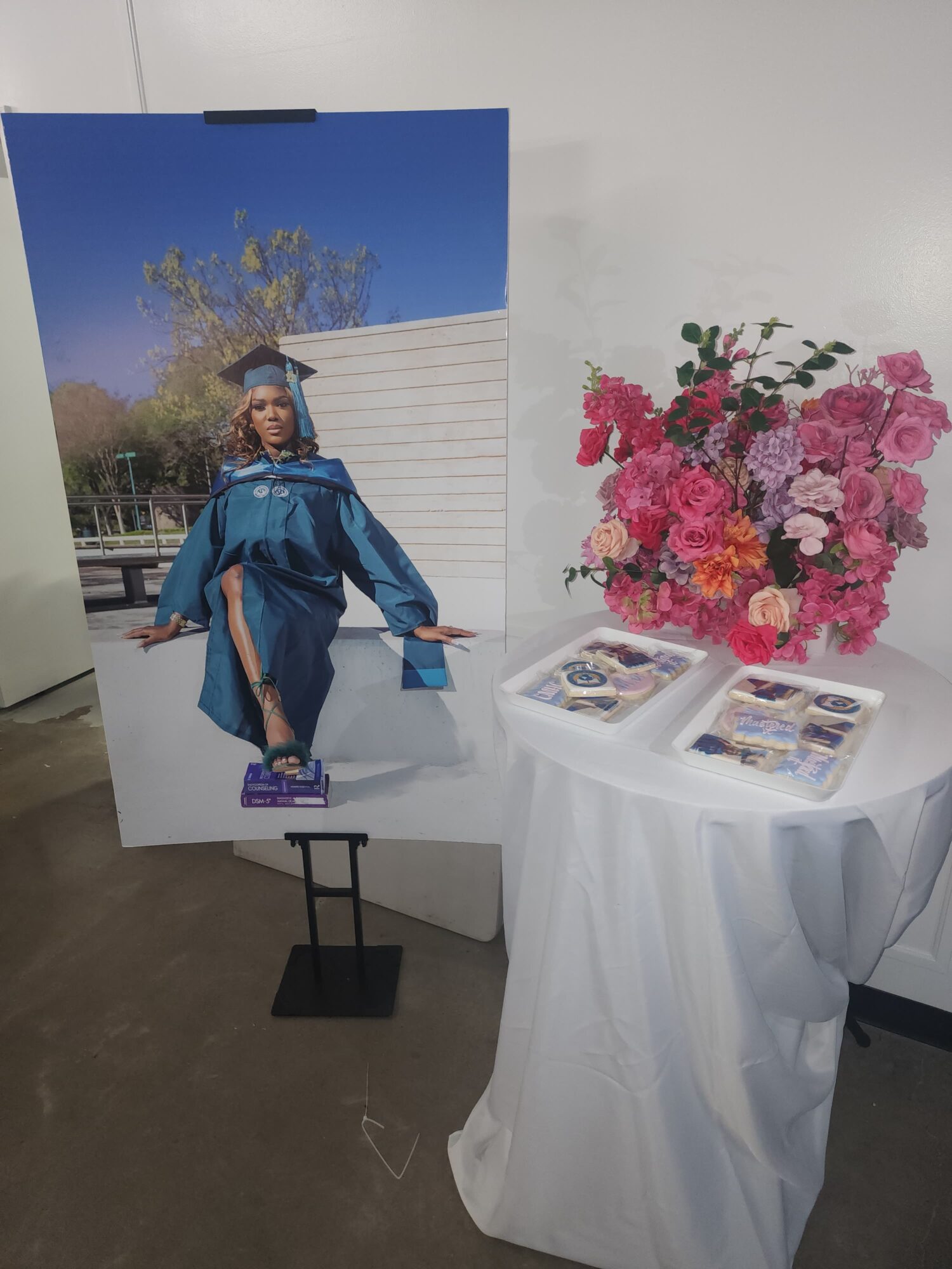 Large graduation photo of a person in cap and gown outdoors, displayed on a stand, next to a table with pink flowers and brochures.