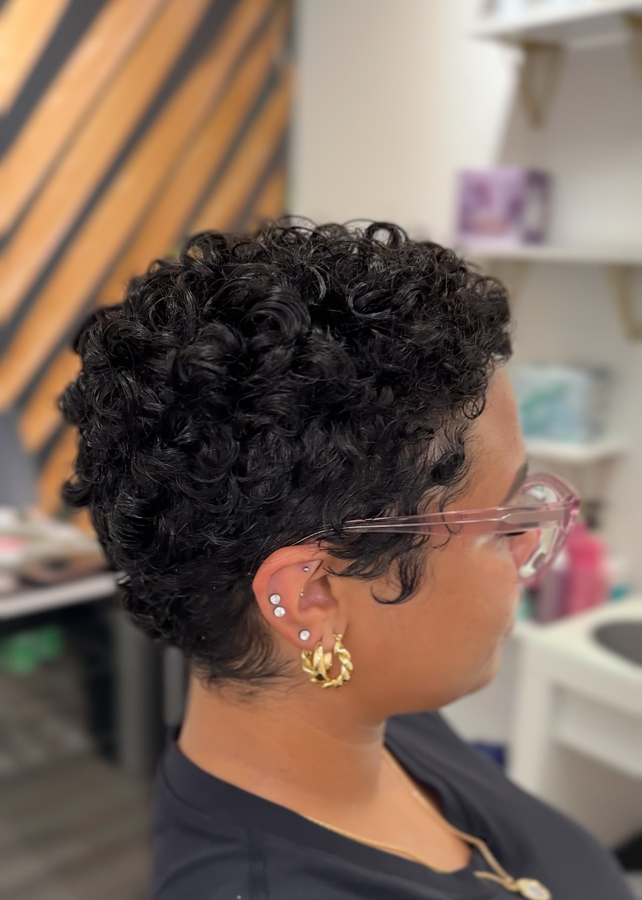 Side view of a person with short, curly black hair, wearing glasses and earrings, indoors with shelves in background.