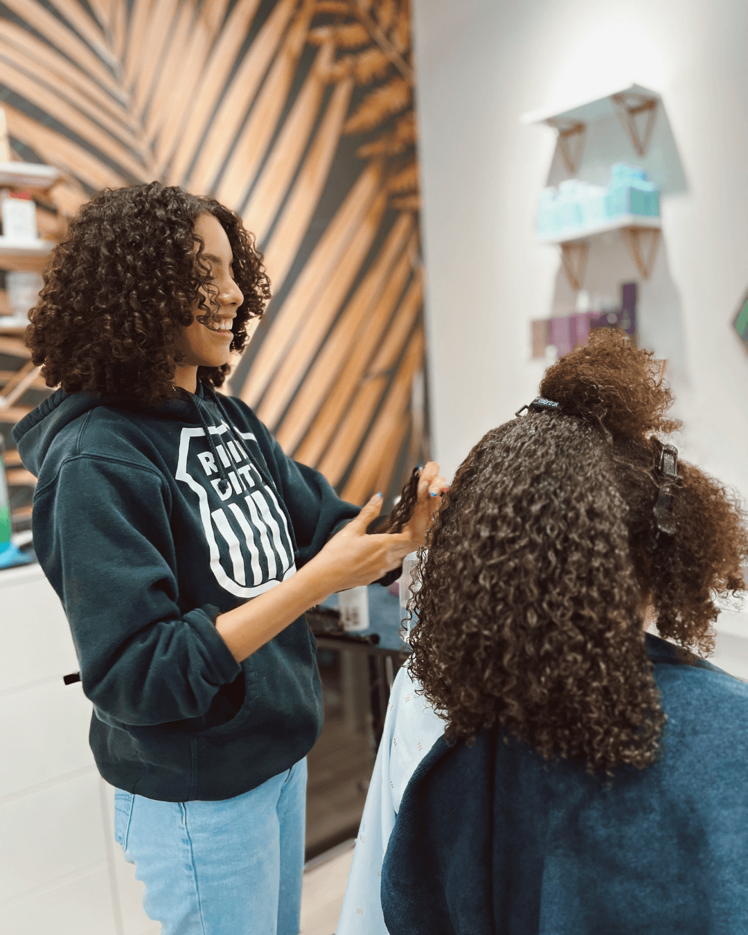 Two women with curly hair in a salon, one smiling and holding a phone, the other with her back to the camera.