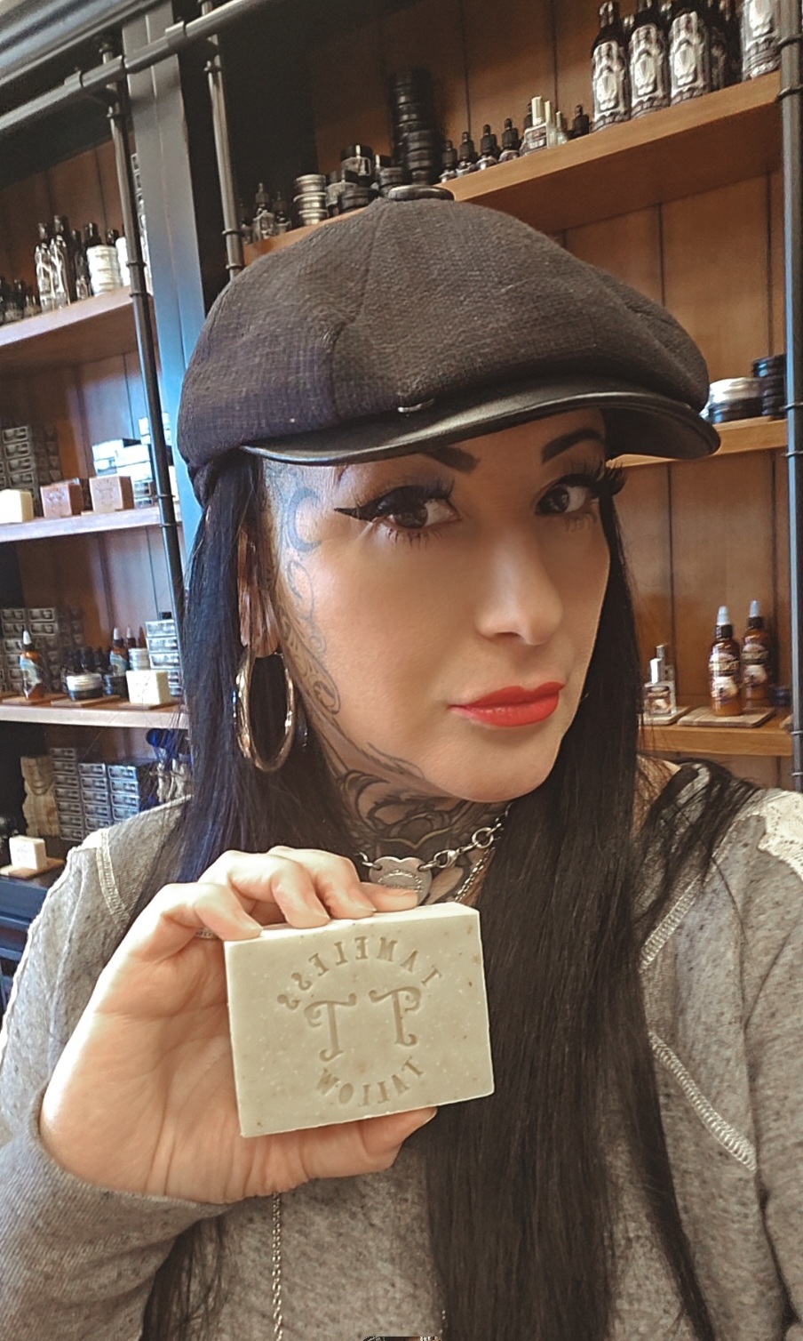 Woman with dark hair, wearing a gray cap and hoop earrings, holding a small rectangular object, in a store with shelves of bottles.