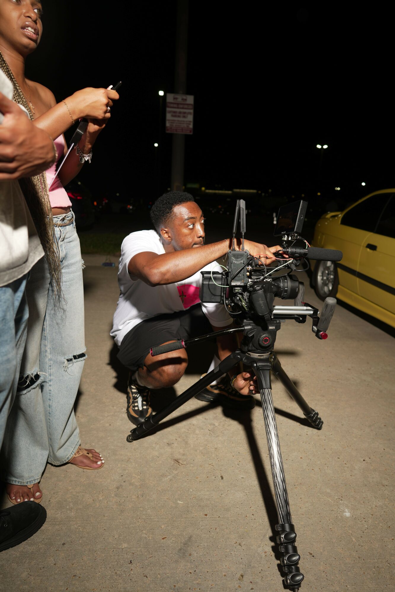 Person in a wheelchair at night, with a person standing nearby, on a street with a yellow vehicle in the background.