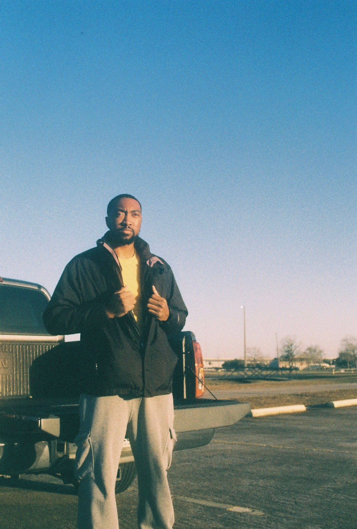 Man standing outdoors near a vehicle, holding a bag, with a clear blue sky background.