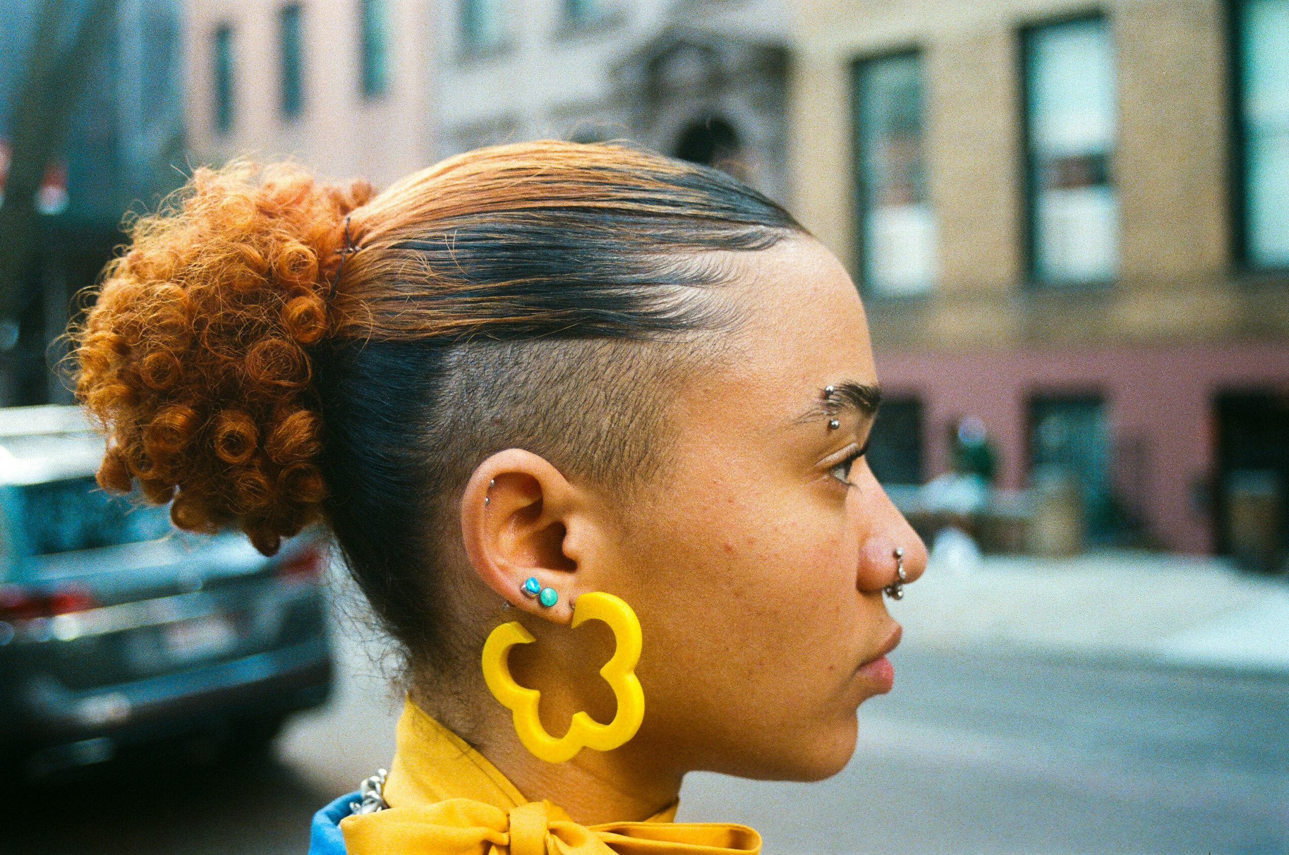 Side profile of a woman with a curly bun and large yellow earrings, outdoors in an urban setting.