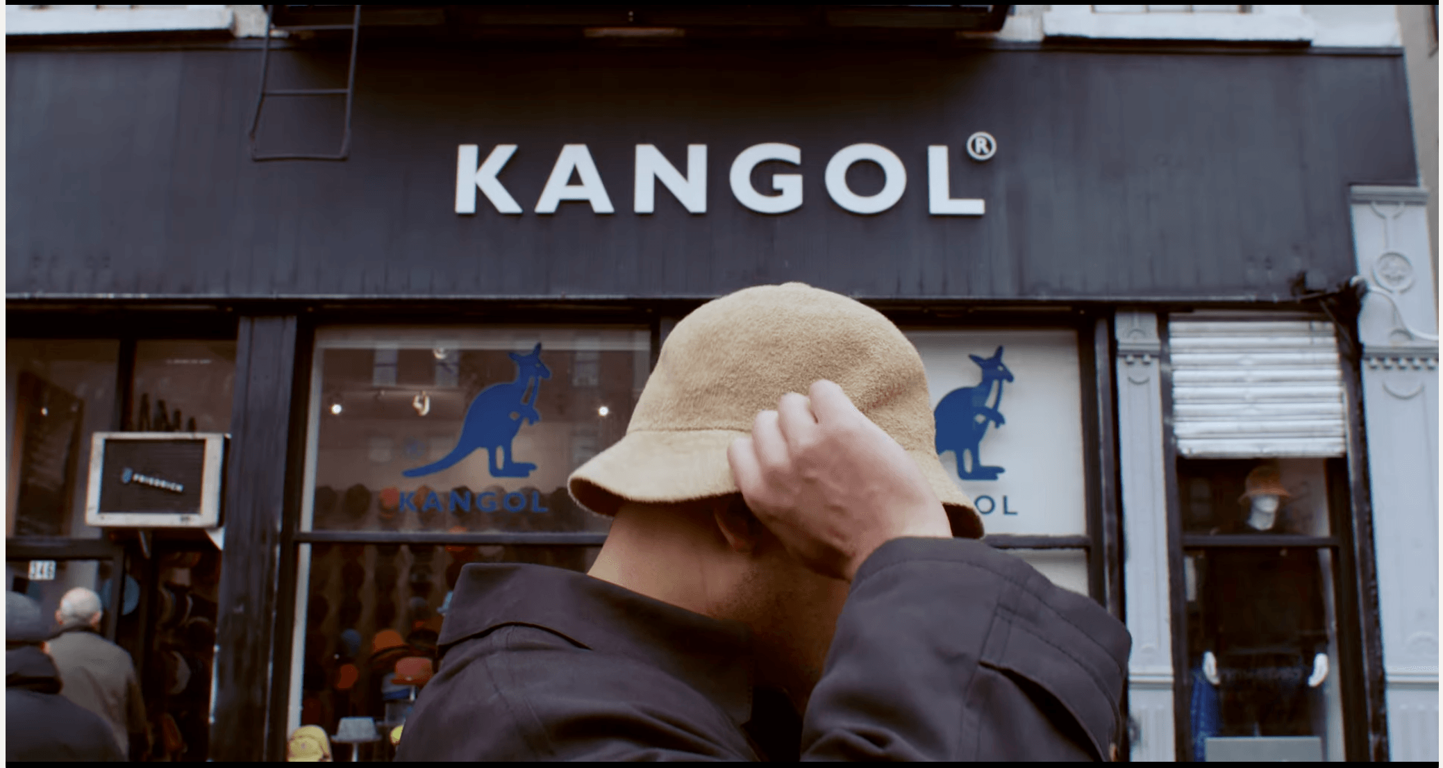 Person wearing a beige hat and dark jacket in front of a store with 'KANGOL' sign, covering their face with hand.