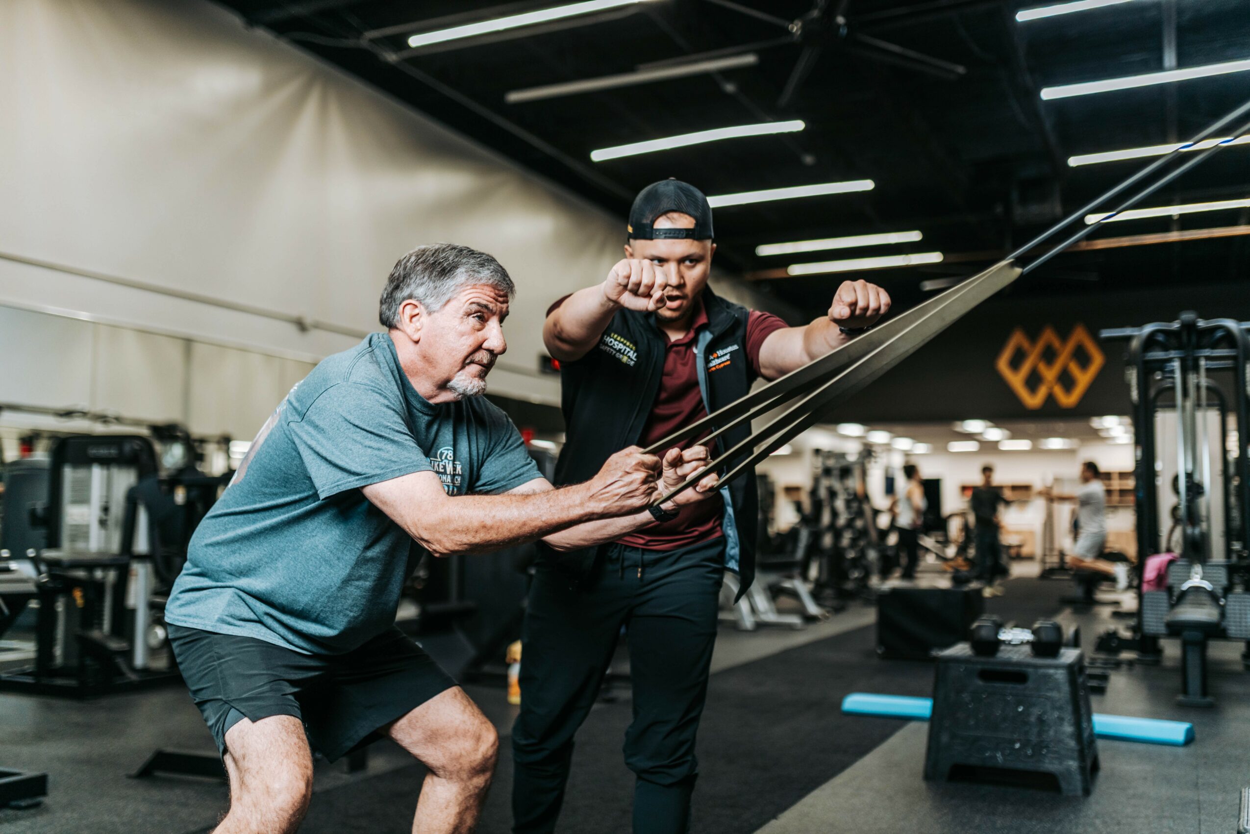 Two men in a gym, one older and one younger, working out together with resistance bands, surrounded by gym equipment.
