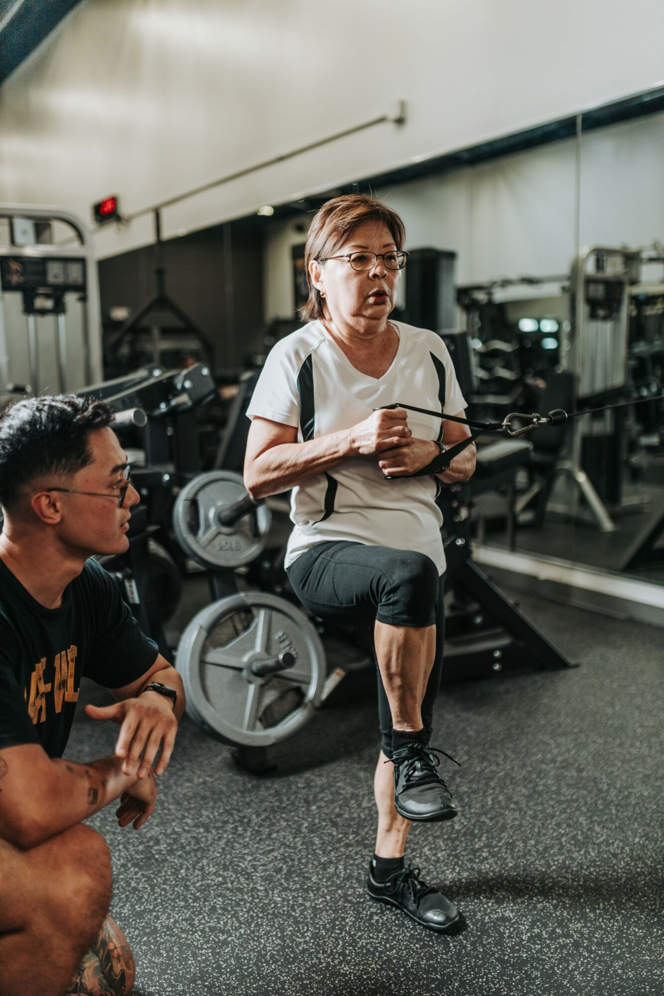Woman instructing a fitness class in a gym, with a man sitting nearby, gym equipment in background.