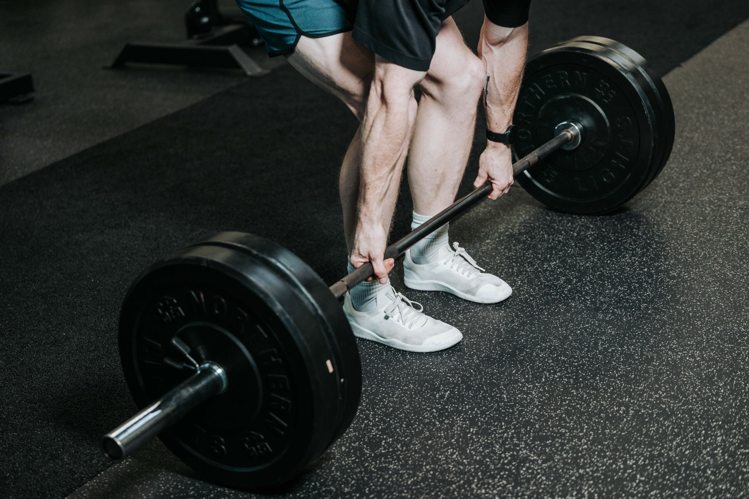 Person lifting a barbell with weights in a gym, wearing white shoes and black shorts.