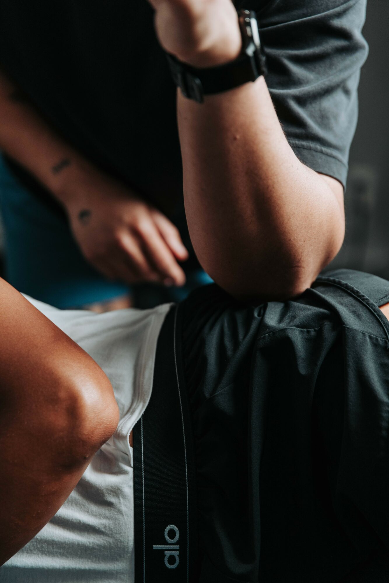 Person resting arm on knee, wearing a watch, with a backpack nearby, in a casual setting.