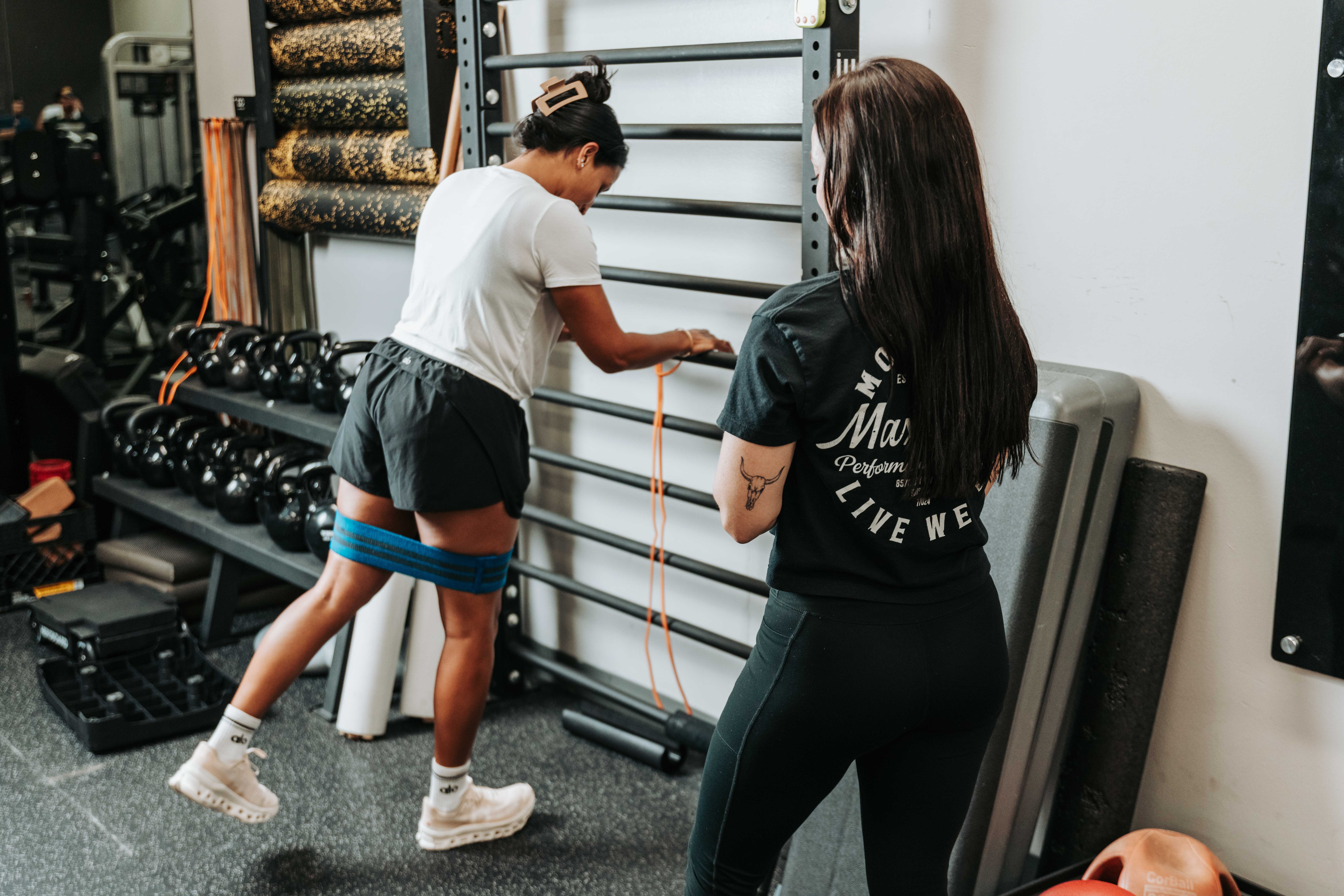 Two women in a gym, one using resistance bands around her legs, the other observing, with gym equipment in background.