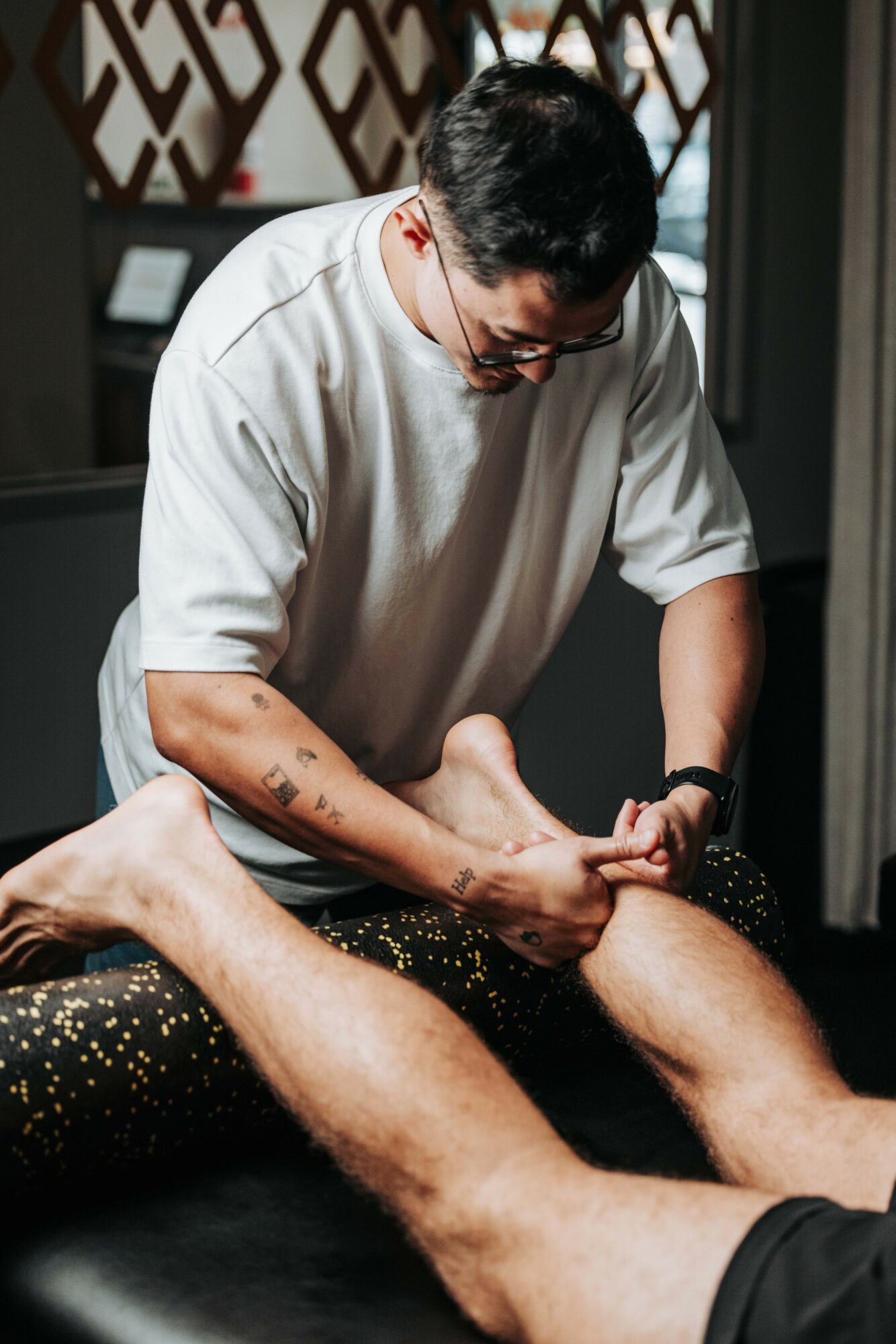 Person receiving massage, focusing on leg, in a room with dark background and patterned wall, wearing glasses and a white shirt.