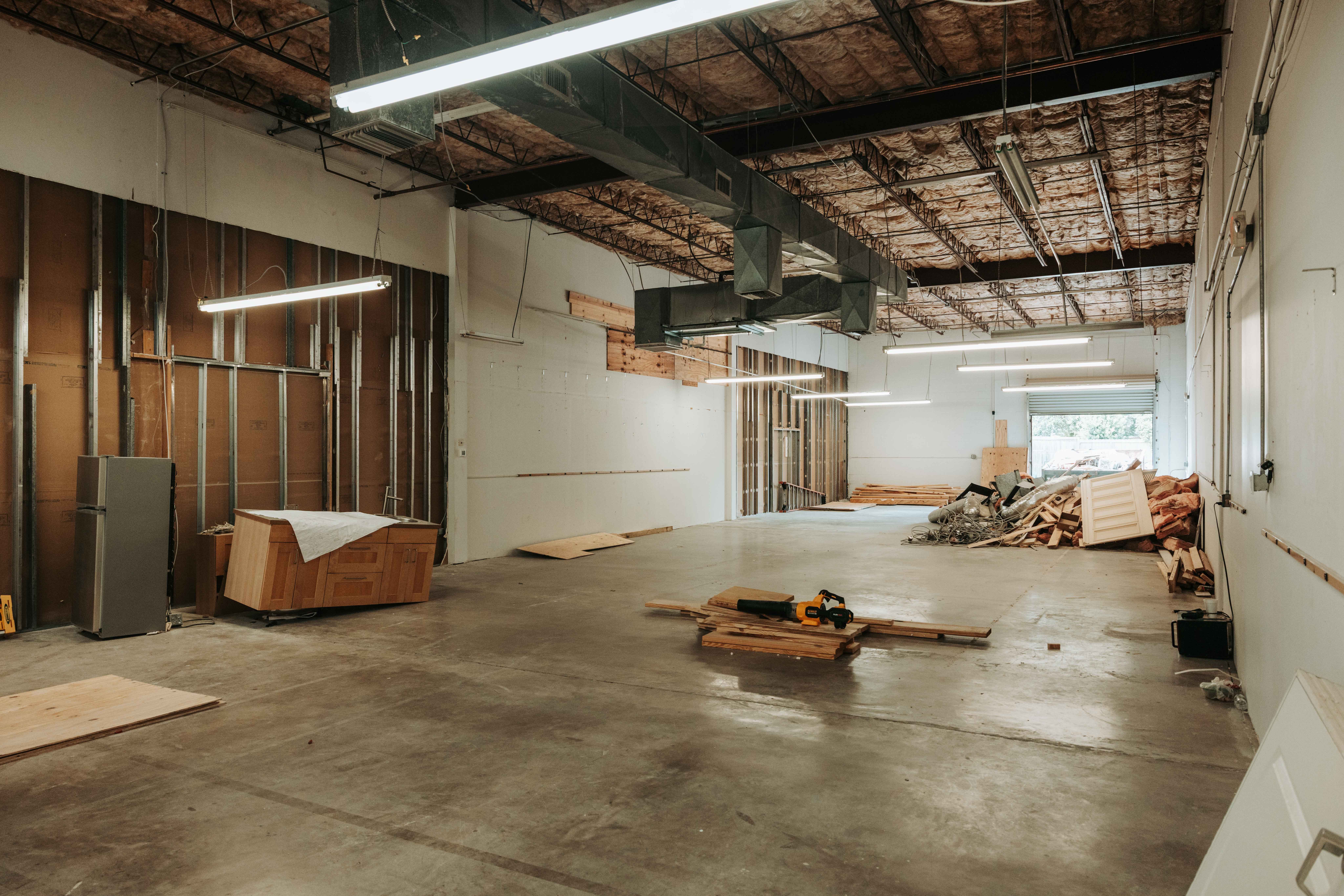 Empty industrial space with exposed ceiling, wooden wall, and scattered construction materials, including a bed frame and pallets.