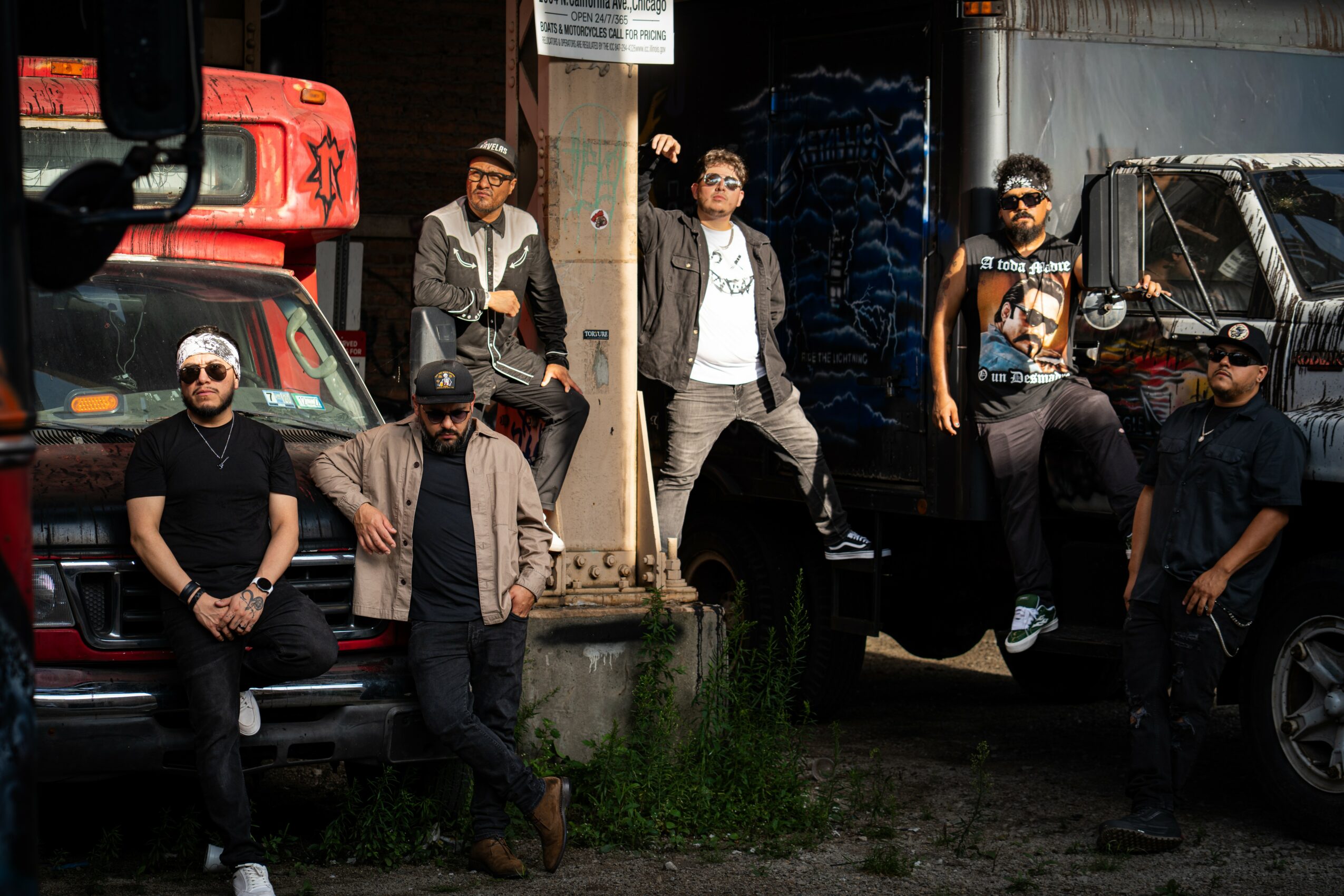 Group of six people standing and sitting near trucks and vehicles outdoors, some wearing sunglasses and casual clothing.
