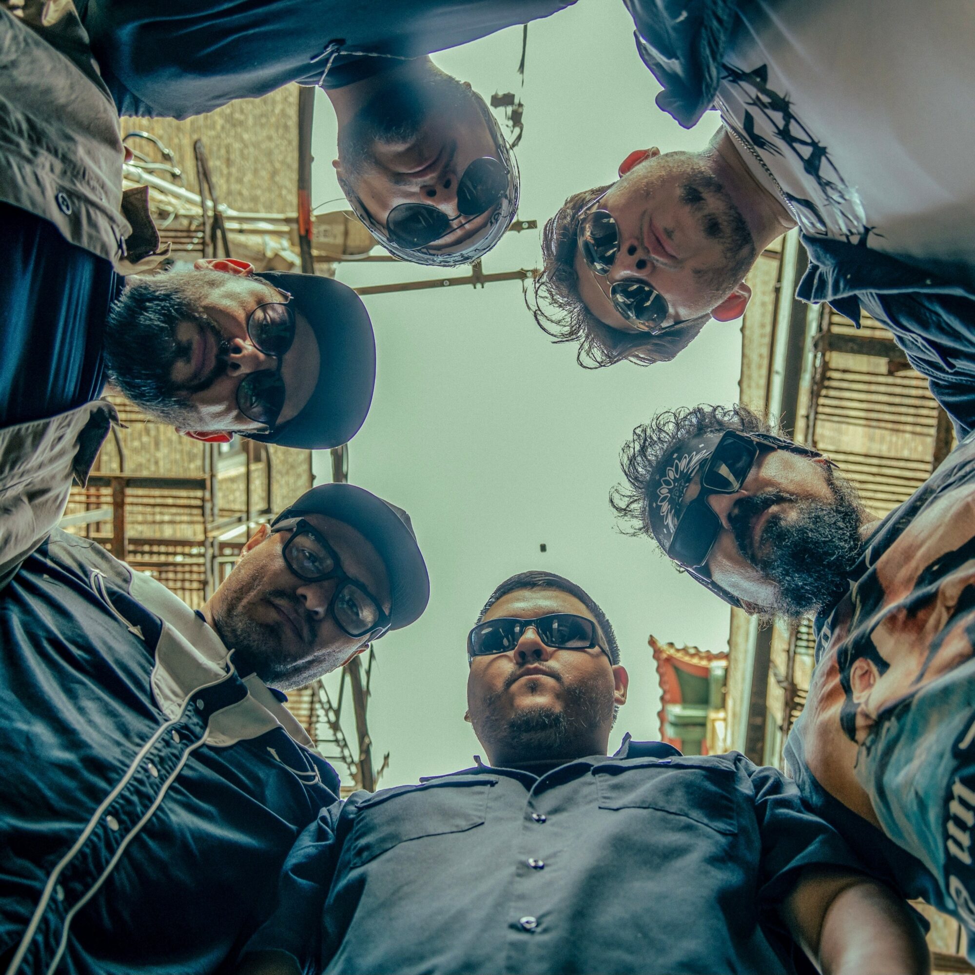 Group of six people looking down at camera, wearing sunglasses, outdoors, with wooden structures in background.