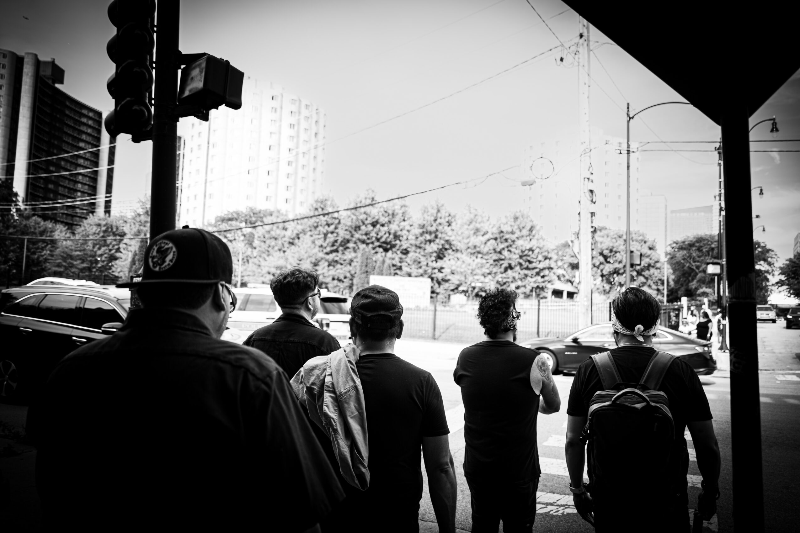 Five people waiting at a crosswalk on a city street, with cars and buildings in the background, in black and white.